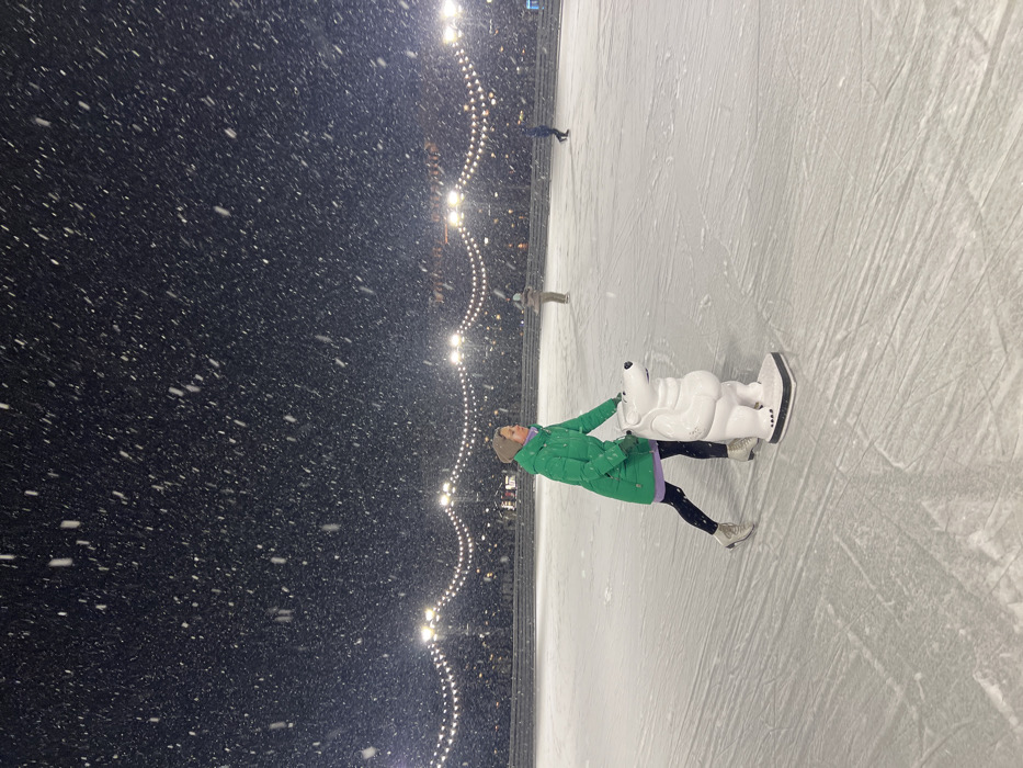 An outdoor ice rink on New Holland Island, Saint Petersburg, Russia, is depicted at night during heavy snowfall. A female individual, wearing a bright green winter coat, black leggings, white ice skates, and a light-colored beanie, is positioned slightly left of center. She is actively ice skating, her left leg extended behind her, while holding onto a white, dog-shaped ice skating aid with both hands. A purple layer is visible beneath her green coat. Her gaze is directed towards the right side of the frame. In the mid-ground and background, two other individuals are also seen ice skating. The ice surface shows visible skate marks. Overhead, strings of illuminated lights are strung between poles, providing illumination against the dark sky, which is filled with numerous falling snowflakes. Structures or tiered seating are vaguely visible beyond the rink perimeter in the background.