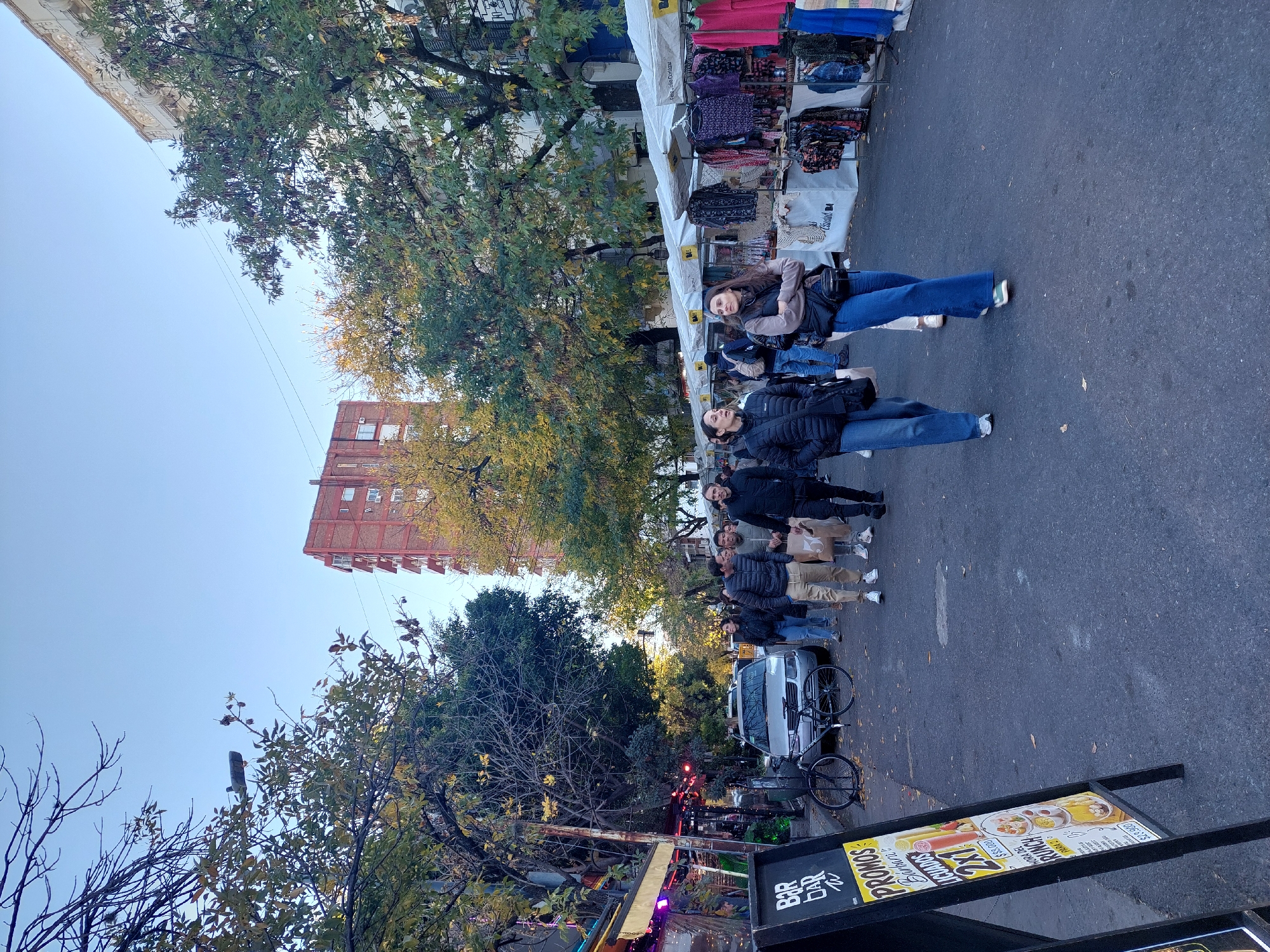 The image depicts a street scene in Buenos Aires, Argentina, specifically in the neighborhood of Palermo, likely during a weekend outdoor market. A row of people, primarily adults, are walking along a street lined with stalls displaying merchandise, presumably clothing or accessories. They appear to be engaged in shopping or browsing. Some are carrying shopping bags.
A silver van and a bicycle are parked alongside the market stalls. Trees line the street, some with yellowing leaves indicating autumn. A tall brick building is visible in the background.
In the foreground, a sign advertising food and drinks is partially visible. The sign features the text "Bar Bar" and "PROMOS LICUADOS 2x1", suggesting it advertises a nearby bar or restaurant with special offers on smoothies.
