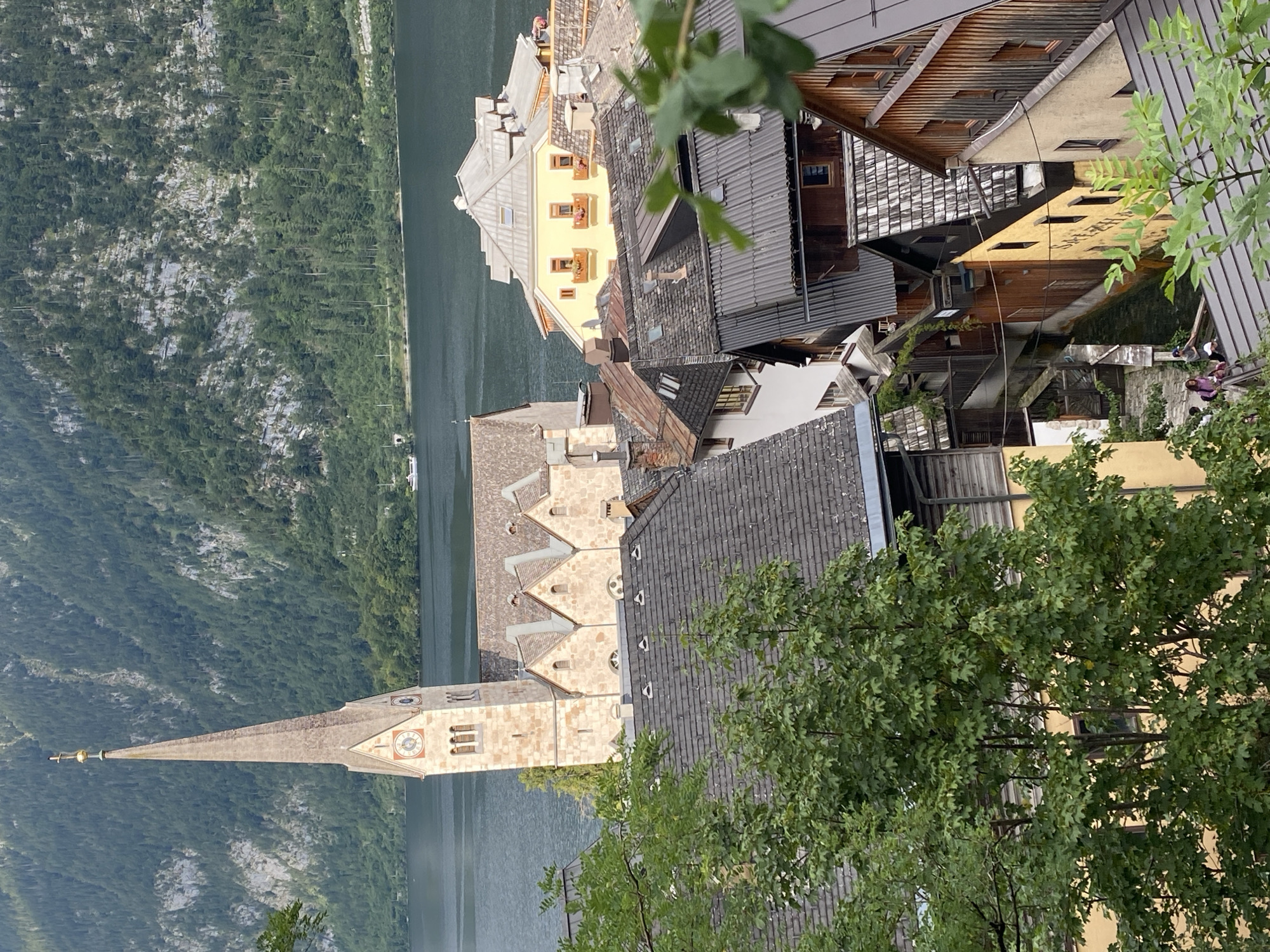 This image captures a scenic view of Hallstatt, Austria, dominated by the town's iconic Pfarrkirche (Parish Church) with its tall, slender spire. The church, constructed of light-colored stone, features a clock on its tower and is flanked by a Gothic-style building with a tiled roof. Below the church, a cluster of traditional Austrian houses with steeply pitched, dark tiled roofs are densely packed, with some featuring visible wooden elements and chimneys.
The town is situated on the shore of a dark, calm lake, which occupies the middle ground of the image. In the background, a steep, densely forested mountain rises, its slopes covered with lush green trees and rocky outcrops. A small white boat is visible on the lake, indicating calm water conditions.
In the foreground, several large green trees with visible leaves obscure parts of the lower section of the image. Within the narrow, winding streets or pathways between the houses, two individuals are discernible. They appear to be walking or standing on a bridge or elevated walkway, interacting casually, with one person wearing a pink shirt and the other a gray shirt.
The architecture of Hallstatt is characterized by its close proximity and integration with the natural landscape, with houses built on steep inclines and narrow plots of land. The overall atmosphere is one of a charming, historic European village nestled within a dramatic natural setting.