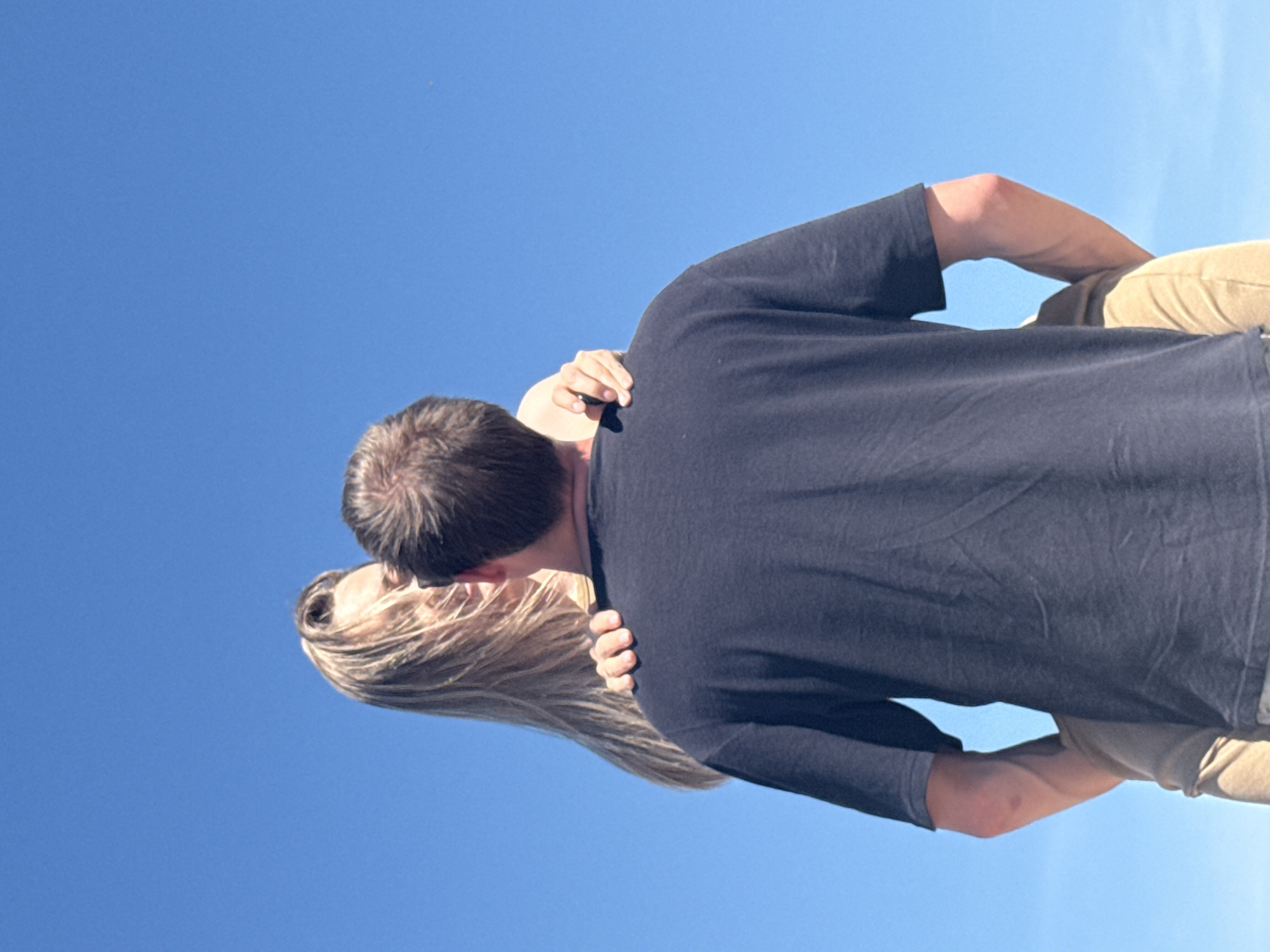 A couple is embracing under a clear blue sky. The woman has blonde hair, and the man has short brown hair and is wearing a dark blue T-shirt and khaki pants. The man has his arms around the woman. The location is not discernible as Chaikovsky, Russia from the image.