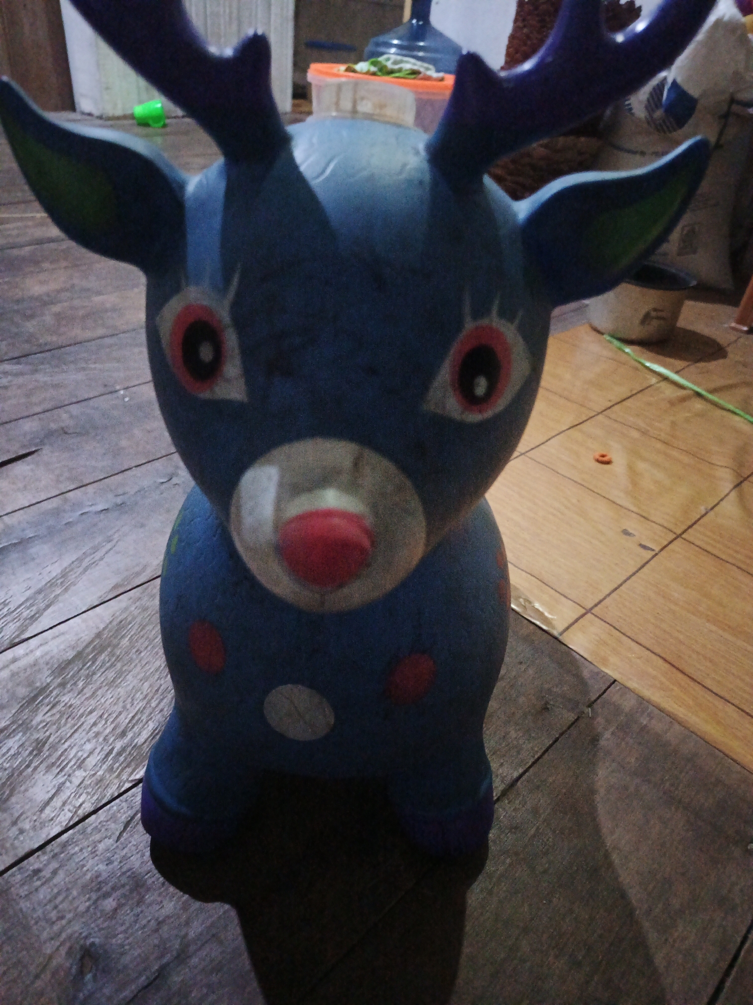 A blue, inflatable reindeer toy with purple antlers sits on a wooden floor. The toy has large, circular eyes with red and black irises, and a red nose. It is decorated with red and white polka dots. The background is blurred and shows a white wall, a blue fan, and some sacks. The floor appears to be made of wooden planks. The scene is dimly lit. The location is not specified within Sinar Bulan, Indonesia.