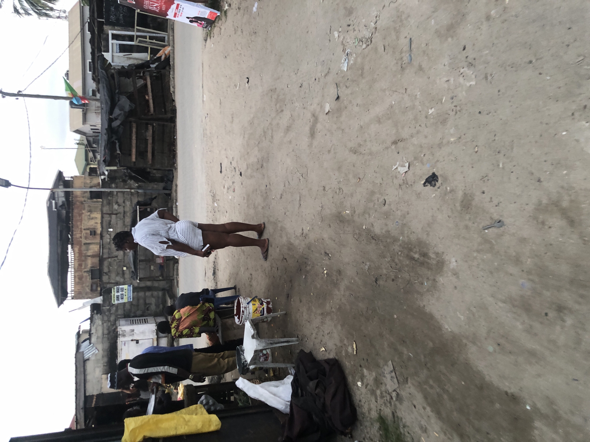 In Ado, Nigeria, a woman wearing a striped shirt and shorts stands with her back to the camera on a dusty road. She is holding a phone in her right hand. Several people are visible in the background, near a structure that appears to be a roadside stall. One person, possibly a vendor, is handing something to another person. A few individuals are seated on stools or chairs, and there is a large bucket near a white stool. Buildings made of brick and corrugated metal are visible behind them, along with some wooden structures covered with tarps. A sign that reads "JAMB WAEC NECO GCE" is attached to a brick wall. Further down the road, a large white poster with red lettering is partially visible. The ground is covered in sand and debris, with some patchy vegetation. The sky is overcast.