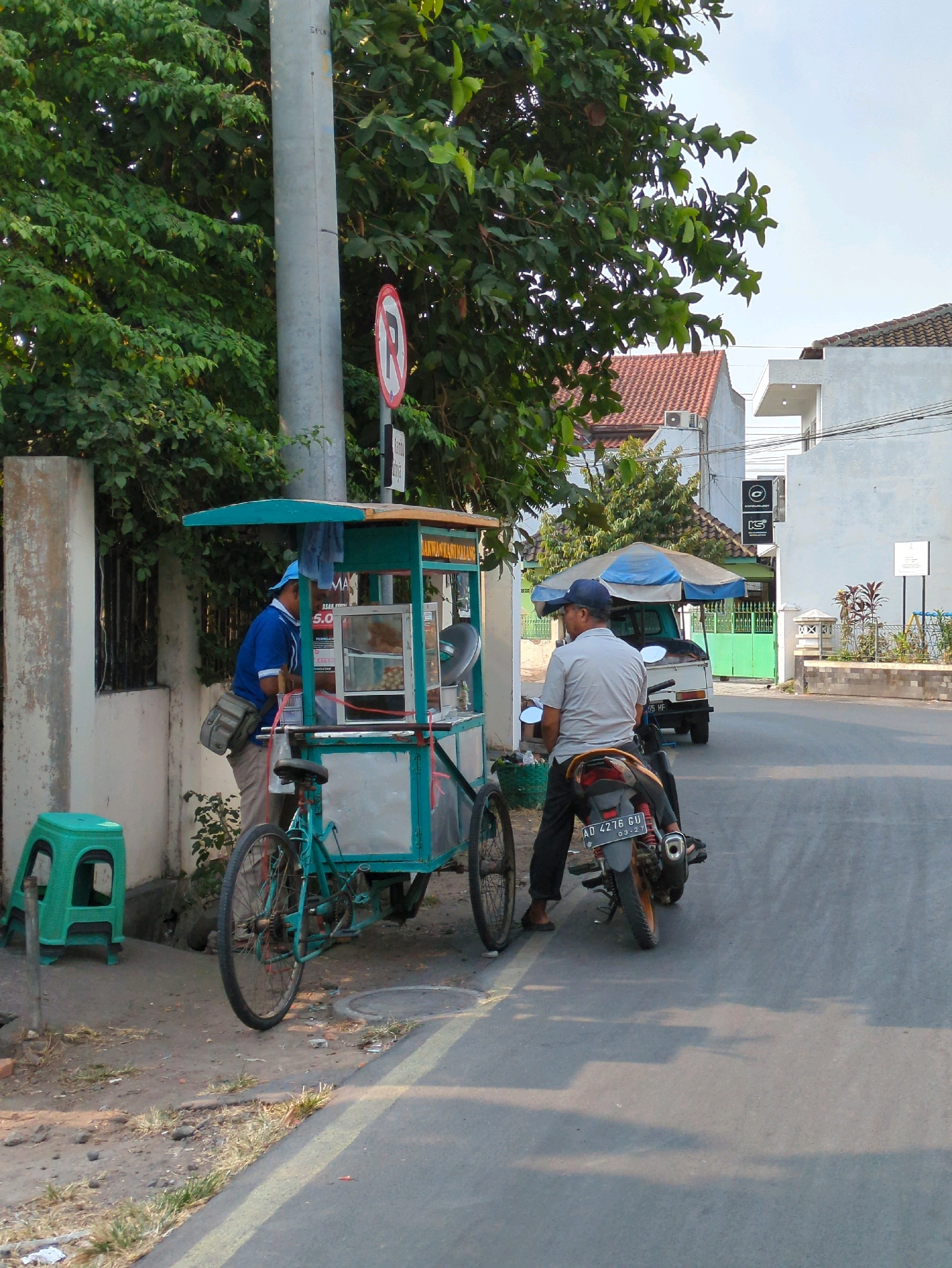 In Surakarta, Indonesia, a street scene unfolds with a bicycle cart vendor and customers. A vendor, wearing a blue shirt and a beige bag across his chest, stands beside his turquoise and green cart, which appears to be selling food. Next to the cart, a bright green plastic stool is visible.
A customer, wearing a light gray polo shirt and a dark blue cap, sits on a motorcycle in the street, facing away from the cart. The motorcycle has a license plate that reads "AD 4276 GU". The cart is a three-wheeled vehicle, with one wheel visible at the front.
Behind the vendor and cart, a tall metal pole stands with a circular red and white sign bearing the letter "R" inside a red circle, indicating a no-entry or restricted zone. Lush green trees and vegetation are abundant in the background. Buildings with tiled roofs and white walls are also present. The street is paved with asphalt and marked with a yellow line. The sky is clear and blue. The overall impression is a snapshot of daily life and commerce on a street in Surakarta.