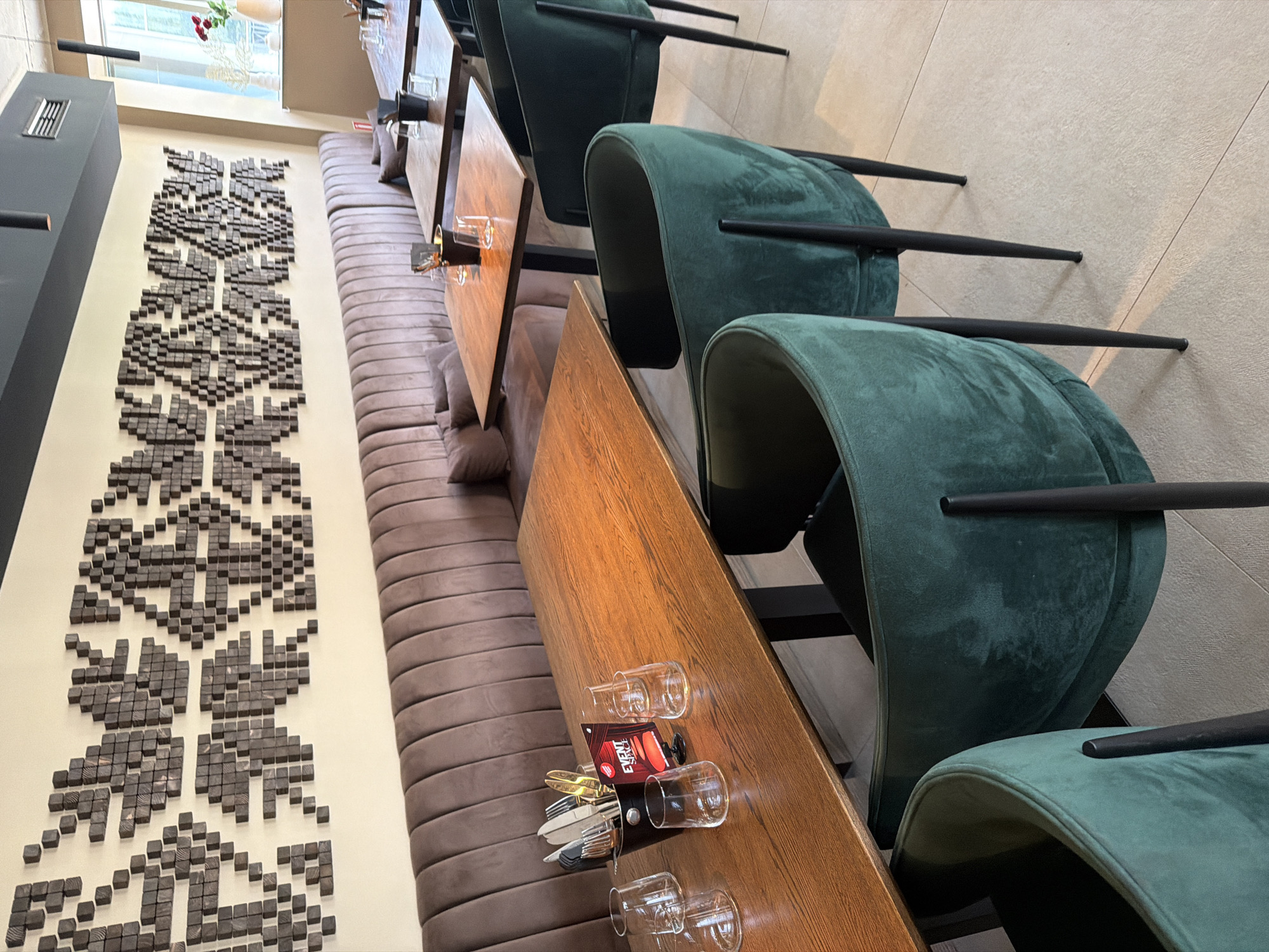 This image depicts the interior of a modern restaurant or cafe located in Sofia, Bulgaria. The scene presents a well-lit dining area devoid of people, suggesting it is unoccupied.
The primary visual elements include a long banquette of taupe-colored, vertically ribbed upholstery that stretches along the left wall, accompanied by individual dark green velvet dining chairs with slender black legs. Multiple wooden tables, primarily rectangular, are set for dining. The table in the immediate foreground features a black bucket containing an assortment of silverware, including some gold-toned utensils, and four clear, stemless drinking glasses. A small tabletop sign, partially readable as "EVENT! SPACE", is also present on this table. Further tables in the midground are similarly set.
Above the banquette, a distinctive wall decoration covers a significant portion of the wall. This artwork consists of numerous small, dark wooden blocks arranged to form large, pixelated geometric patterns that resemble traditional textile designs. The ceiling is dark grey or black, featuring integrated lighting fixtures, cylindrical pendant lights, and an HVAC vent. To the right, a large window allows natural light into the space, and a small arrangement of red flowers in a vase is visible on a windowsill. The floor is covered with light beige or off-white tiles.
