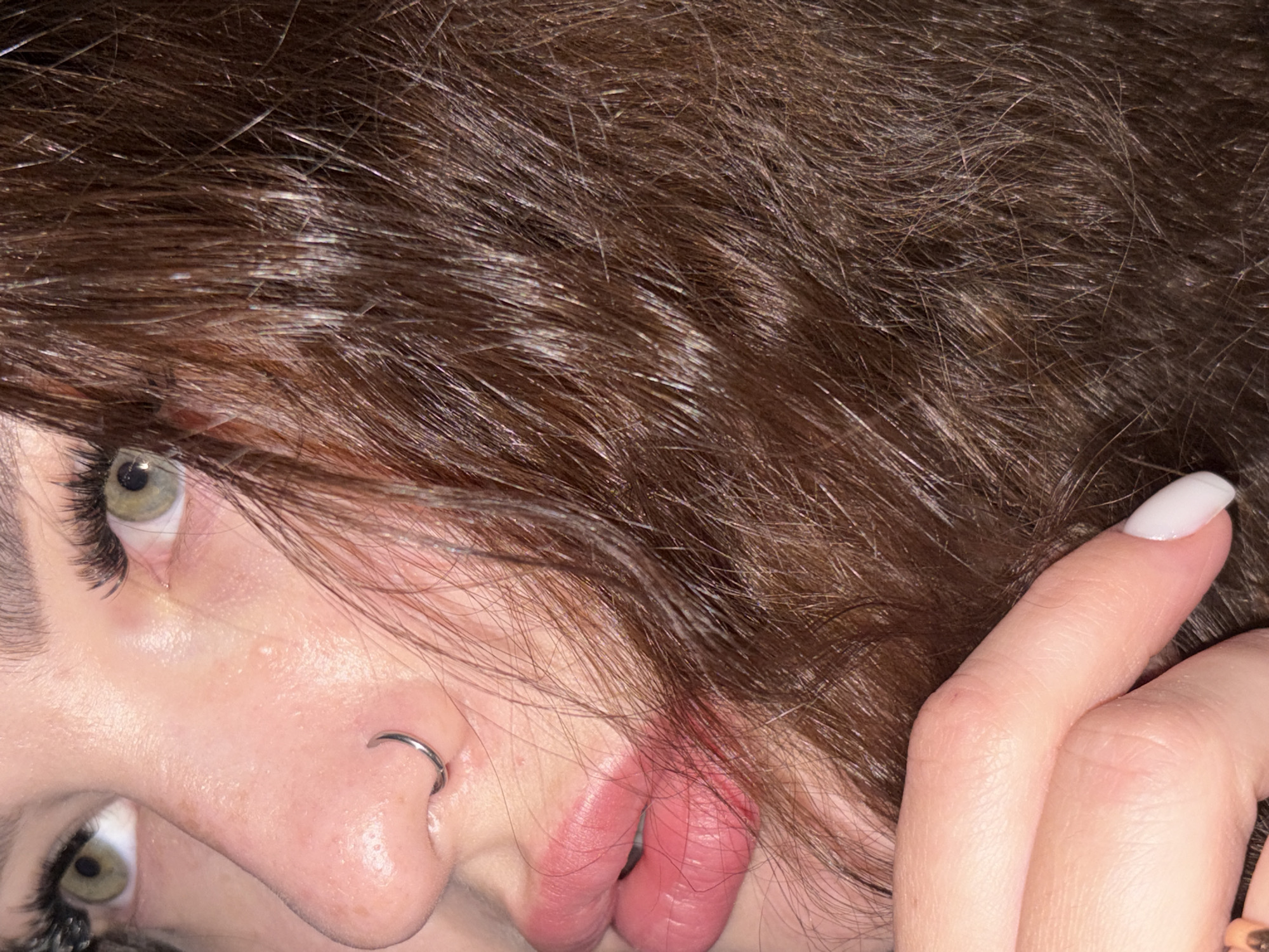 A close-up image shows a person's face and hand. The person has light skin, green eyes, and dark brown hair. The person is wearing a nose ring in the left nostril. Their lips are slightly parted. Their hand is near their face, with a white nail polish on the finger closest to the camera. The hair is in the foreground. There is no context provided to suggest a location in Krasnodar, Russia.
