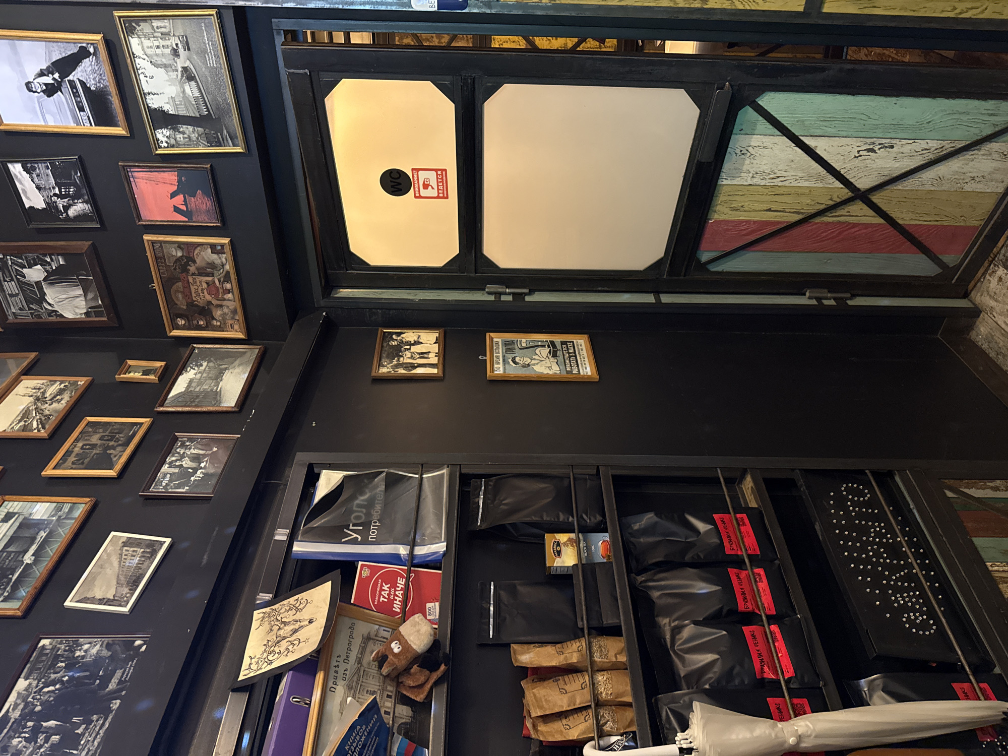 The image depicts the interior of a cafe or shop, possibly a coffee shop, with a rustic and vintage aesthetic. The walls are painted black and adorned with numerous framed photographs and art prints, mostly in black and white, some with sepia tones, depicting historical scenes, portraits, and street views.
On the left side, there is a shelf unit filled with various items. These include packaged coffee or tea bags, some with red labels indicating "БРАЗИЛИЯ РЕЛИК" (Brazil Relic), and other unmarked black bags. A white umbrella is leaning against the shelf. Below, a black rectangular object with many small, light-colored circular elements arranged in a pattern is visible. There are also books and a stuffed toy horse on higher shelves.
On the right side of the image, there is a dark-colored door, possibly leading to a restroom, with two rectangular panels. The upper panel has a "WC" sign and a red square sticker with white text that appears to be a warning or informational notice. The lower panel is a lighter color. The door is framed by colorful wooden planks arranged vertically and diagonally, creating a patchwork effect.
The overall atmosphere suggests a place with a focus on historical decor and possibly artisanal products. The presence of "Saint Petersburg" in the prompt, combined with the style of photography and lettering, suggests a location in that city.