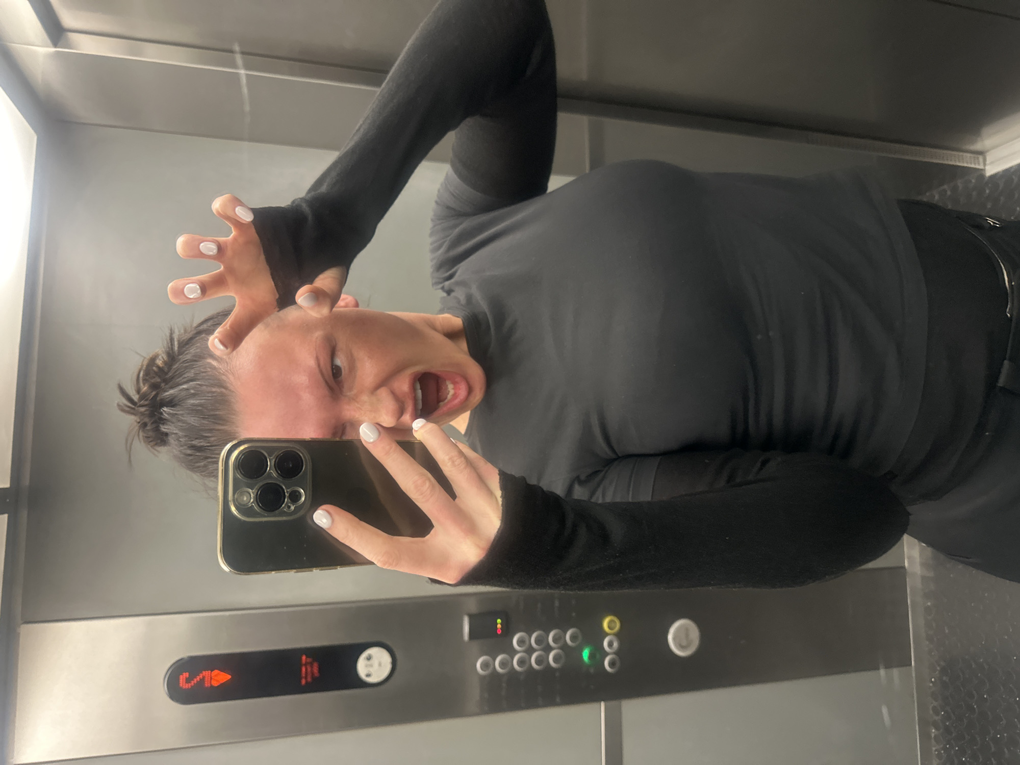 The image depicts a young adult female inside an elevator, taking a selfie using a smartphone. She is positioned centrally, facing a reflective surface which captures her reflection. Her right hand, with light-colored nail polish, holds a gold-colored smartphone with a three-lens camera array, obscuring the right side of her face. Her left hand, also with light-colored nail polish, is raised near her head, fingers splayed in a "claw" gesture. Her mouth is open in a wide expression, consistent with a shout or roar. Her hair is styled in a bun. She wears a black, long-sleeved top, possibly athletic wear, featuring thumbholes.
The elevator interior features brushed gray metal walls. To the left of the subject, a dark gray elevator control panel is visible. This panel includes a red digital display showing an illuminated "S" with an upward arrow, signifying a floor destination or current location (likely "Service" or "Sub-basement") and ascent. Below this, a column of circular silver-colored buttons represents floor selections. Further down, two green illuminated buttons and two yellow illuminated buttons are present, with a circular silver emergency call button at the bottom. The elevator floor is a dark, textured metal, possibly diamond plate.
The elevator's characteristics, specifically the "S" floor indicator commonly denoting "Service" or "Sub-basement" in British English elevator systems, suggest the location is consistent with elevators found in the city of London, United Kingdom. The precise building or address within London cannot be determined from the available visual information.