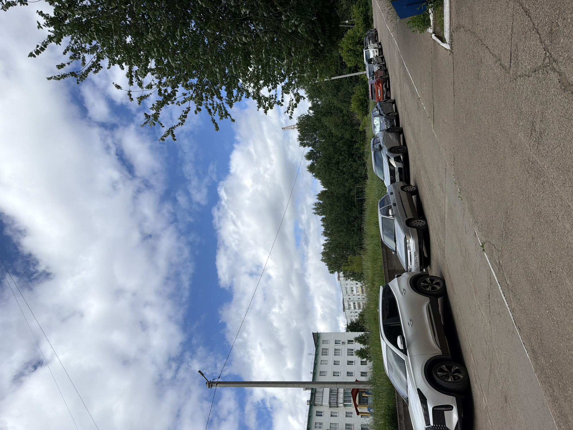 The image shows a row of parked cars on a paved road next to a grassy area. In the background, there are trees and residential buildings. The sky is blue with scattered white clouds. A concrete utility pole with wires is visible on the left side of the image. The scene appears to be an outdoor urban or suburban setting. The time of day seems to be daytime, possibly late morning or afternoon, given the brightness of the sunlight. The weather is partly cloudy. The crops show a more detailed view of the cars and the sky. One of the license plates is partially visible and reads "E587PY 199". The image appears to be taken in Chaikovsky, Russia, as indicated by the location context. No specific activity or event is depicted, other than the cars being parked. There are no people visible in the image.