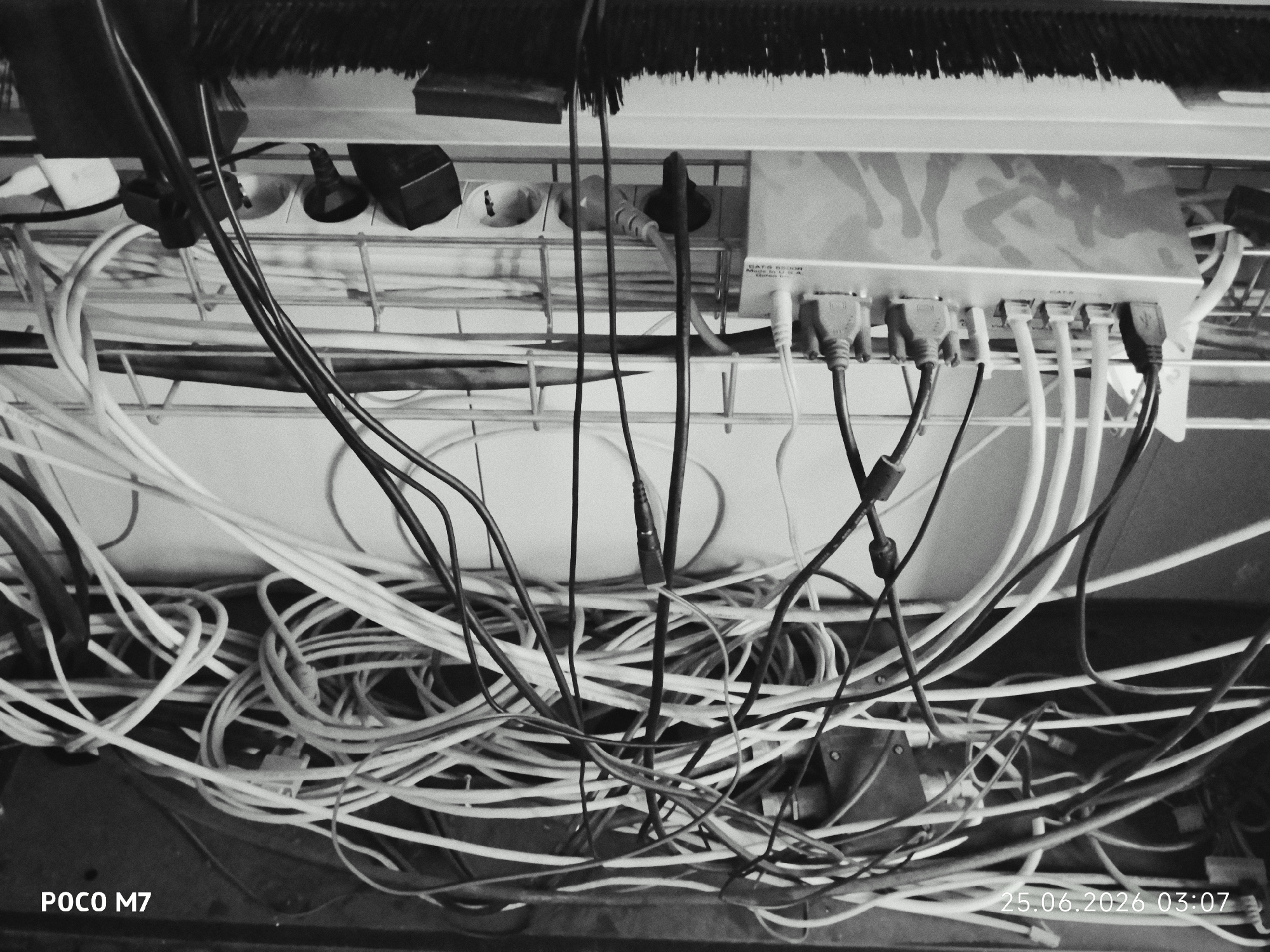 A black and white, high-angle medium shot captures a cluttered collection of electronic cables and a power strip on a metal rack. The scene is set in an indoor environment, possibly a utility closet or behind a desk, with a brush visible at the top of the frame. Several black and white cables are tangled together, some plugged into the power strip, while others hang loosely. A silver-colored device with multiple ports, likely a network switch or hub, is also connected to some of the cables. The image is dated "25.06.2026 03:07" and watermarked "POCO M7". There are no people present.