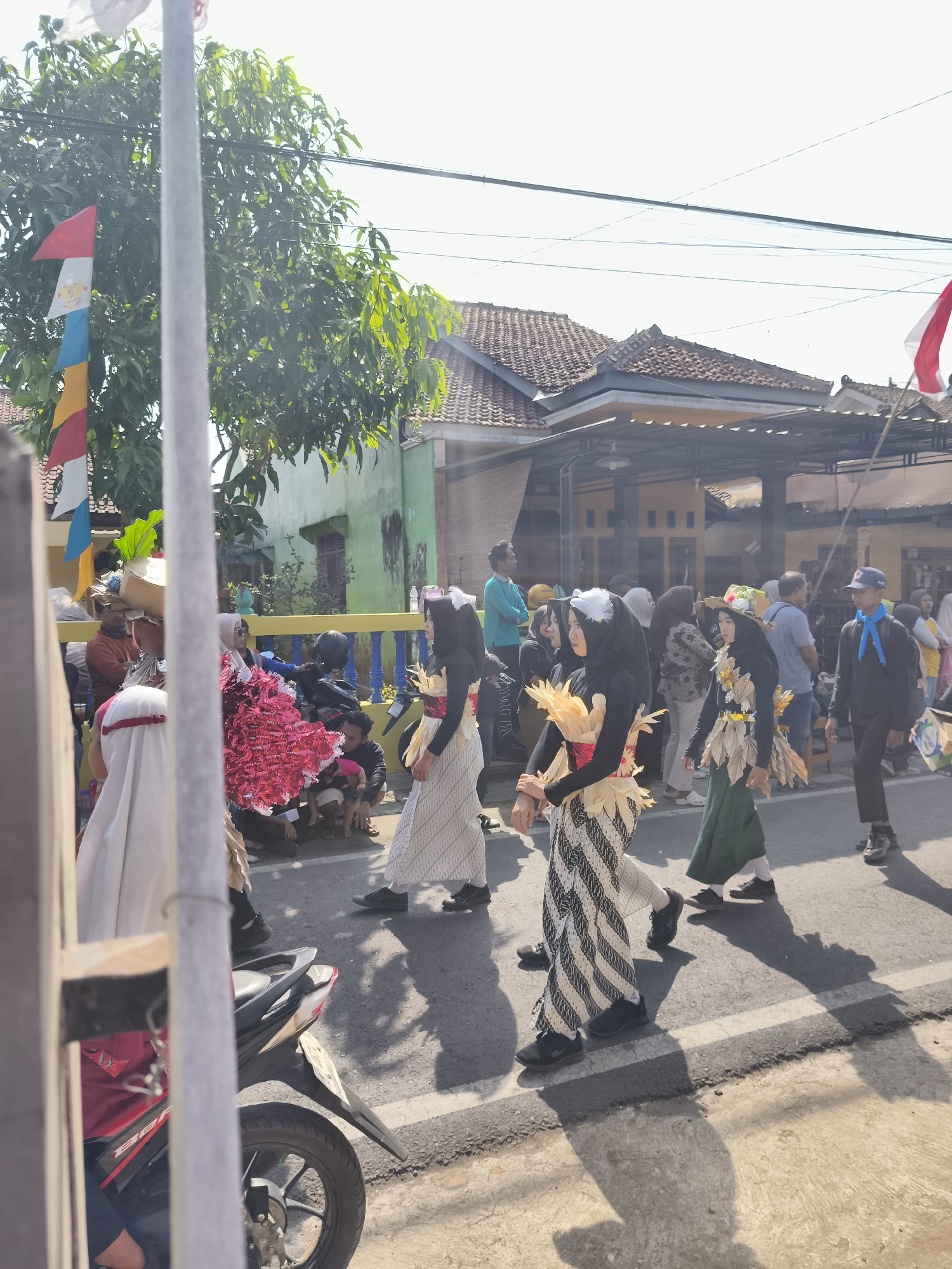 In Kutoarjo, Indonesia, a procession is underway on a paved street under a bright sky. Three young women, dressed in matching black hijabs and distinctive costumes, are walking. Their outfits feature white and black striped skirts or trousers, and their tops are adorned with layered, tan-colored fabric resembling leaves or petals, accented with white decorative elements. One of the women wears a straw hat with similar floral embellishments. Behind them, a crowd of spectators and participants is visible. Notable objects in the scene include a motorcycle parked on the side of the road, several buildings with tiled roofs, and colorful flags. The sunlight casts long shadows of the people onto the street. The event appears to be a cultural parade or celebration.