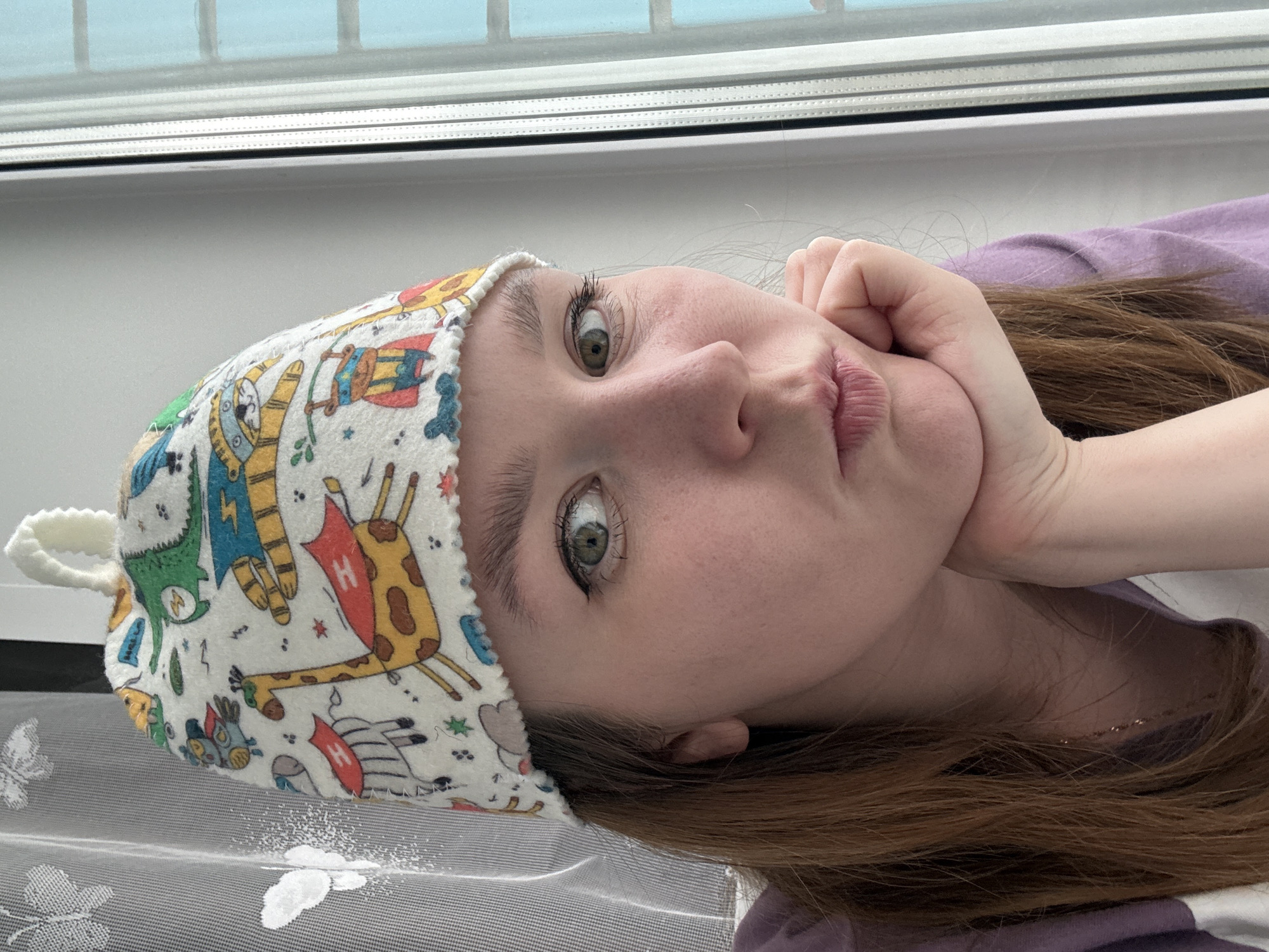 A young adult female is depicted in a close-up indoor shot, understood to be located in Izhevsk, Russia. She has light brown hair, fair skin with visible pores, and greenish-hazel eyes with winged eyeliner and mascara. Her left eye is notably wider open than her right, both gazing directly at the camera. Her lips are slightly parted in a pout, and her chin rests upon her right hand, which is visible. She wears a purple garment, possibly a t-shirt. Adorning her head is a white fleece hat featuring a colorful, scalloped edge and a small loop handle at the top. The hat's pattern displays various anthropomorphic animals dressed as superheroes, including a green dinosaur with a lightning bolt, a zebra and a giraffe with red capes marked with an "H", and striped animals (possibly badgers or raccoons) in blue masks. The background on the left reveals a translucent white curtain with embossed butterfly patterns, with a gray striped material behind it. On the right, a white window frame with multiple panes is visible, looking out onto a blue, possibly sky-filled, exterior.