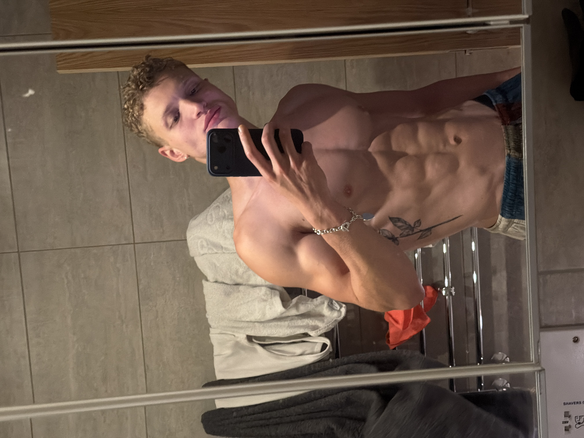 This image, taken in a bathroom in London, United Kingdom, captures a young adult male taking a mirror selfie. The male has short, curly, light brown hair and fair skin. He is shirtless, revealing a muscular physique with visible abdominal muscles and biceps. A silver chain bracelet with a circular charm is worn on his left wrist. A tattoo depicting a plant stem with leaves extends vertically down his left side, below the ribcage. He is holding a dark-colored smartphone in his left hand, partially obscuring his face below the eyes as he gazes towards the camera. He wears patterned shorts, visible from the waist down. The reflection shows light grey tiled walls behind him, along with a vertical towel rail draped with dark and light grey towels. A light-colored garment hangs above the towels. To the right of the reflection, a wooden surface, possibly a door or cabinet, is visible. At the bottom of the reflected scene, a red cloth is seen on a lower rack, and a white panel with partial text "SHAVERS O" and electrical symbols, consistent with a shaver socket, is also visible.