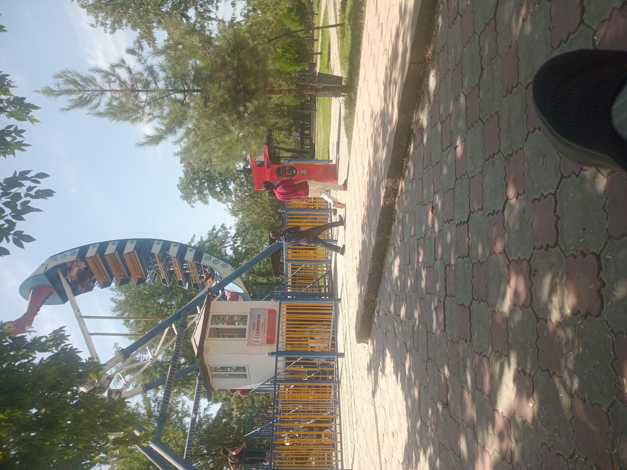 In an outdoor amusement park, a Ferris wheel with blue and yellow carriages dominates the background. A small white ticket booth is situated at the base of the Ferris wheel, with a sign displaying "10 SOMONI". Two individuals are seen walking on a paved pathway in front of the ticket booth. One person is wearing a dark shirt and light pants, while the other is in a red shirt and white pants. The pathway is made of interlocking square tiles in red and grey patterns, with shadows cast from overhead trees. To the right of the pathway, there is a green grassy area with several pine trees and a large green trash bin. In the foreground, a person's shoe is visible, suggesting they are seated or standing. The scene is set under a clear blue sky with some scattered clouds, indicating a sunny day. The overall environment suggests a recreational area in a city.