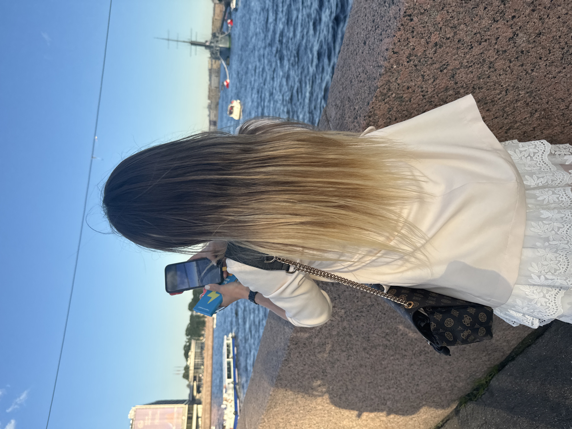 A woman with long, ombre hair stands facing away from the camera, holding a smartphone in her right hand and a blue object in her left, as if taking a photo or video. She wears a cream-colored blazer over a white lace dress, and a patterned handbag with a gold chain strap hangs from her left shoulder. The scene appears to be a waterfront promenade in Saint Petersburg, Russia, possibly near the Palace Embankment, given the presence of a large body of water with boats, including a tall-masted ship in the background and a passenger boat closer to the shore. The sky is clear and blue, indicating a sunny day. The ground in the foreground is a granite walkway.