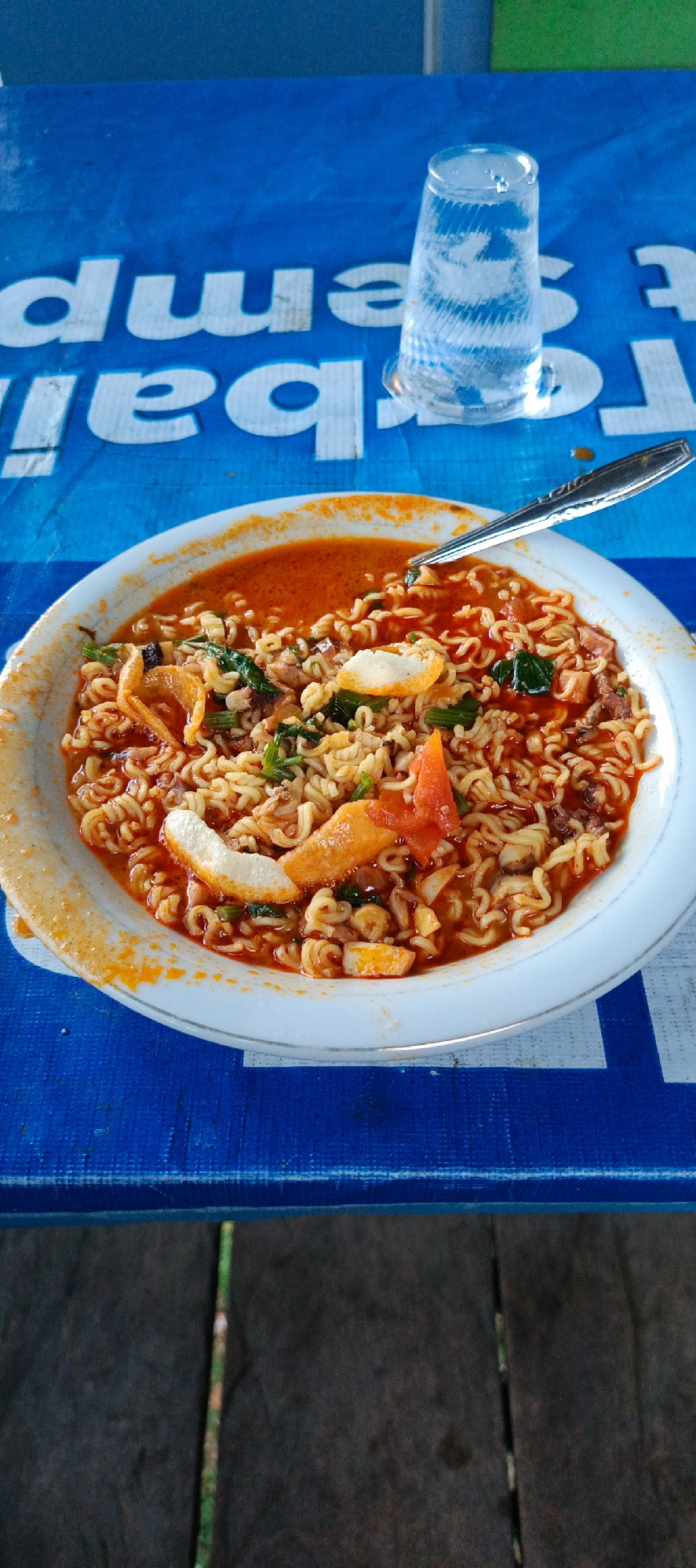 A white bowl filled with instant noodles in a red, spicy broth sits on a blue tablecloth with white lettering. A silver spoon rests in the bowl. To the right of the bowl, a clear glass filled with ice and water is visible. The tablecloth appears to be laid out on a wooden surface.