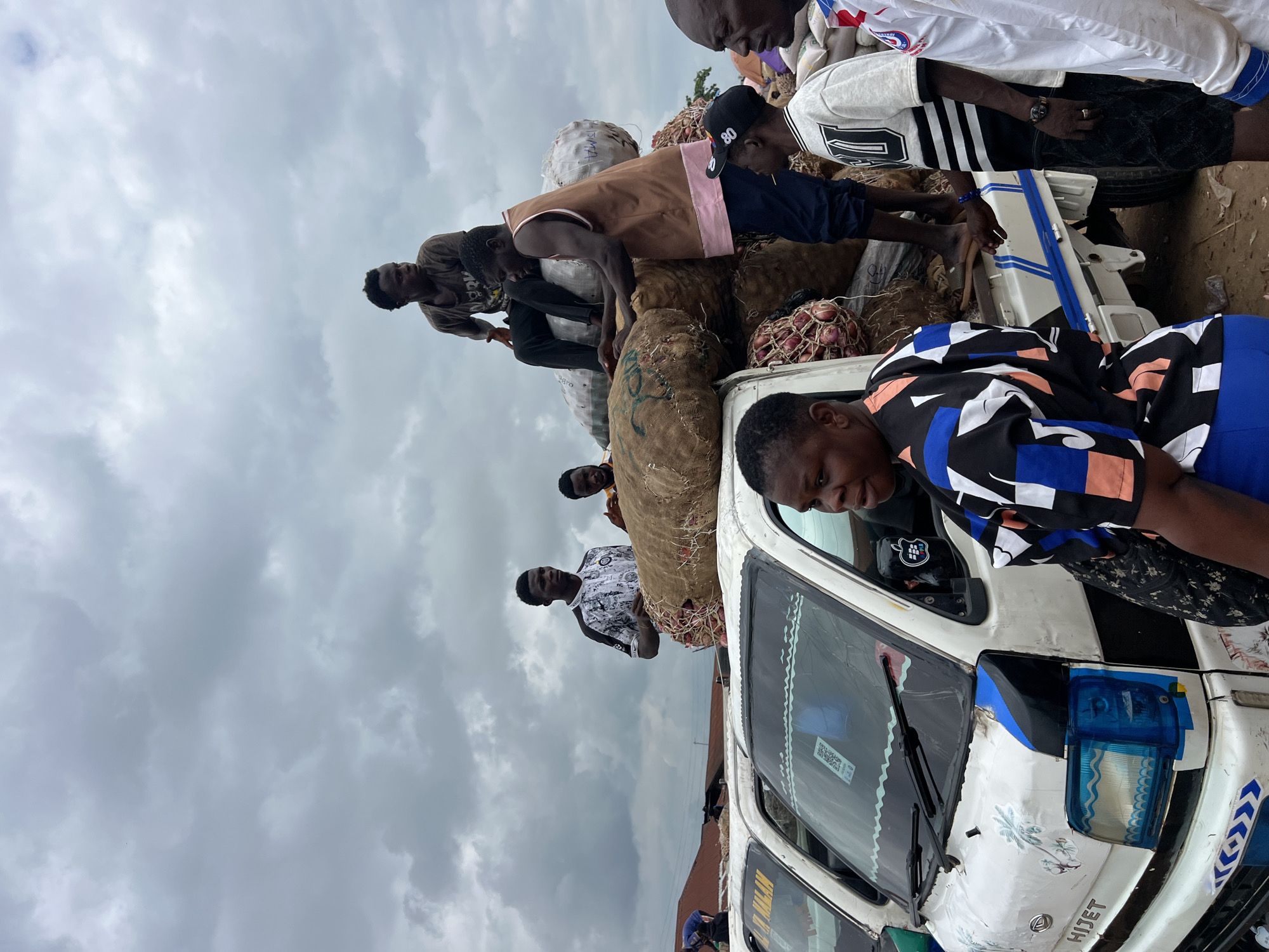 This media file captures a scene of people engaged in loading a van with goods, likely produce, in Oko Amakom, Nigeria. The primary subjects are a group of men, most of whom are actively involved in the process. They are interacting with large sacks and bundles that appear to contain vegetables or other food items, some of which are tied with string.
The scene is set outdoors, with a cloudy sky dominating the background, suggesting it might be overcast or nearing rain. The van itself is white, with visible blue and white stripes along its side. The word "HIJET" is clearly visible on the front of the van. There are also some decorative stickers on the van, including images of palm trees.
The people are dressed in casual clothing. One man wears a brown vest over a dark shirt, another a black baseball cap with "80" on it, and another a white shirt with black stripes. A person in the foreground wears a brightly colored, patterned shirt with blue sleeves. The general atmosphere appears to be one of work and effort, with individuals focused on their tasks.
There are no clear indications of the specific time of day, but the cloudy weather suggests it could be anytime during daylight hours. The location cues, such as "Oko Amakom, Nigeria" and the type of vehicle, help contextualize the scene within a specific region. The activity is clearly related to transportation of goods, possibly to a market or distribution point. No strong emotions are immediately evident, but the focus on the task suggests a practical, work-oriented mood.