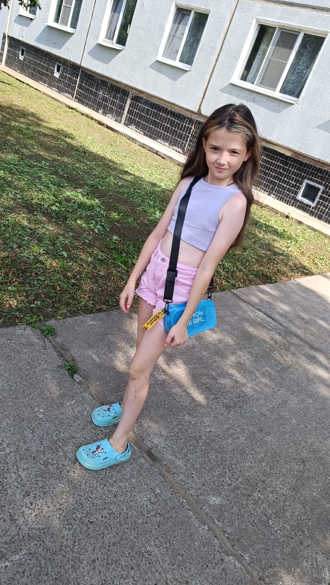 In Chaikovsky, Russia, a young girl with long brown hair, wearing a light purple crop top, pink shorts, and light blue Crocs with Mickey Mouse designs, stands on a concrete sidewalk. She is holding a bright blue cross-body bag with a black strap. The bag has text on it that reads "FASHION GIRL". Behind her, there is a green grassy area and a light-colored building with windows. The building's facade is textured and has dark square tile accents along the lower portion. The scene appears to be outdoors on a sunny day, as indicated by the shadows on the ground.