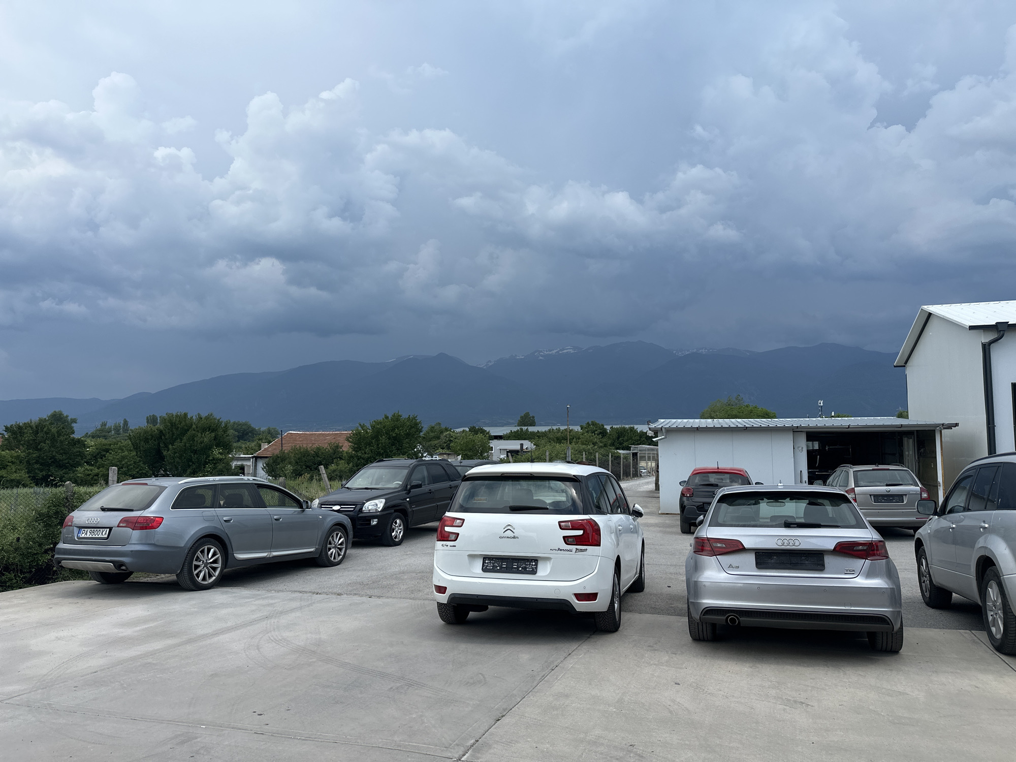 A car lot in Piperevo, Bulgaria, is depicted under a dramatic, cloudy sky. Several cars are parked on a concrete surface. In the foreground, a silver Audi A3 hatchback faces away from the camera, with a silver Audi station wagon to its left. Behind the Audi A3, a white Citroen C4 Picasso is parked. Further back, a black Kia Sportage is visible. In the middle ground, a grey Audi station wagon is parked inside an open garage or workshop area, which has a white exterior and a white roof. A portion of a white building with a black gutter is on the right side of the image. In the background, lush green trees and houses are scattered across the landscape, with a mountain range visible on the horizon under the stormy sky. The overall scene suggests a commercial area with a backdrop of natural scenery, under the impending weather.