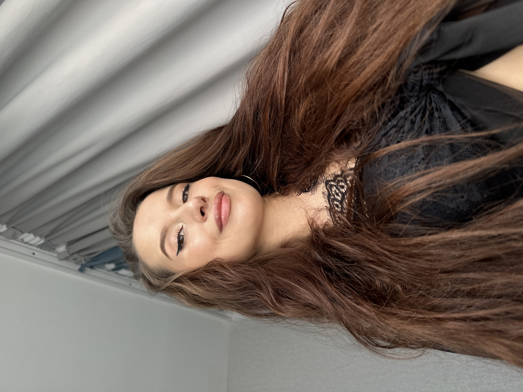 A portrait-style photograph captures a young woman, likely in her 20s or early 30s, in an indoor setting. The image is oriented such that her head is positioned on the left side of the frame, with her body extending to the right, mostly obscured by her hair.
The woman has long, voluminous, dark brown hair that flows dramatically across the majority of the frame, especially to the right, creating a sense of movement and framing her face. Her complexion is clear, and she has visible makeup, including precise winged eyeliner, defined eyebrows, and light pink lip gloss or lipstick on her full lips, which are parted in a gentle, subtle smile. Her eyes are open and gaze slightly upwards and to the right. A small, delicate golden hoop earring is visible on her left ear.
She is wearing a black garment, possibly a top or a bra, featuring intricate black lace detailing visible around her chest and shoulder area.
The background consists of two main elements. On the top left, there is a light grey or white textured surface, possibly a curtain or blinds with vertical folds, suggesting a window nearby. Below this, on the bottom left, is a plain light grey wall. The lighting is soft and even, indicating an indoor environment during daytime, with no direct harsh shadows.
The overall mood of the image is calm and composed, with a touch of allure. There is no visible text in the image. No specific elements within the photograph directly indicate the mentioned location of Yekaterinburg, Russia.