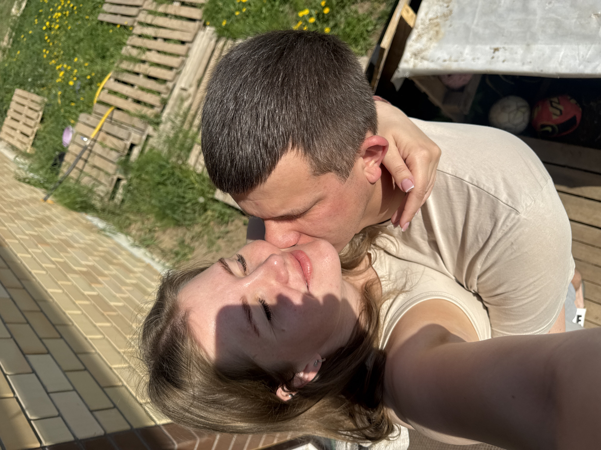 A man and a woman are kissing outdoors. The woman is tilting her head back. The man has short dark hair. She has lighter colored hair. They are wearing light colored t-shirts. The woman is taking the picture. There are wooden pallets and yellow flowers in the background, along with a brick walkway. There are soccer balls under a tarp behind the couple. This photo may have been taken in Ферма, Russia.
