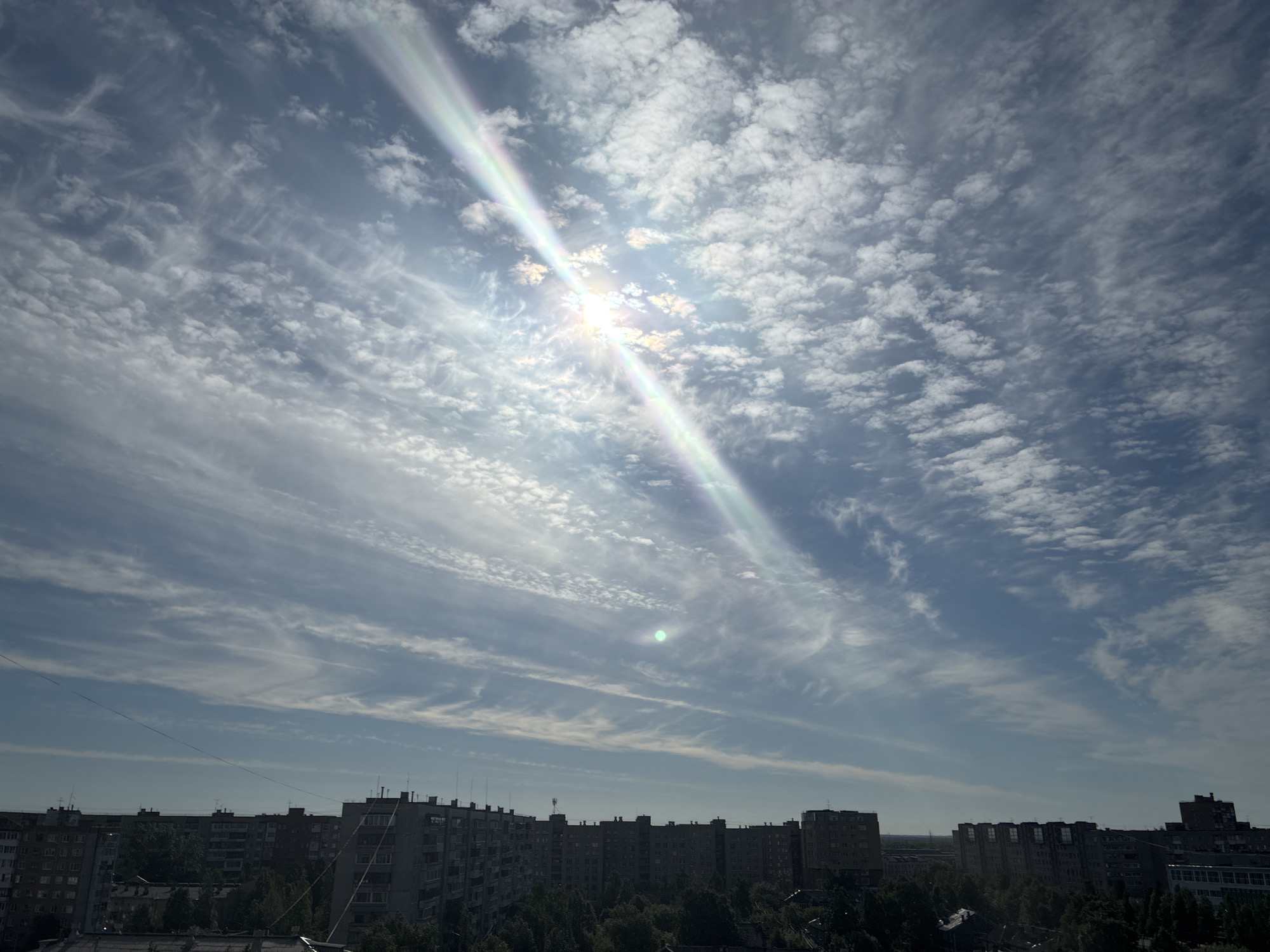 The image is a daytime, eye-level exterior shot of a cityscape under a sky filled with dynamic clouds and bright sunlight. The sun is positioned towards the upper left quadrant of the image, casting a rainbow-like lens flare diagonally across the sky. The clouds are varied, with patches of fluffy cumulus clouds mixed with wispy cirrus formations, creating an uneven texture across the blue expanse. Below the sky, a line of apartment buildings stretches across the horizon. These buildings are predominantly grey and appear to be of Soviet-era architecture, with multiple stories and visible windows. Trees are interspersed between and in front of the buildings, their dark green foliage contrasting with the lighter tones of the architecture and sky. A few thin, dark wires are visible spanning across the frame in the middle ground, likely utility or power lines. The overall impression is of a typical urban environment on a bright, partly cloudy day.
