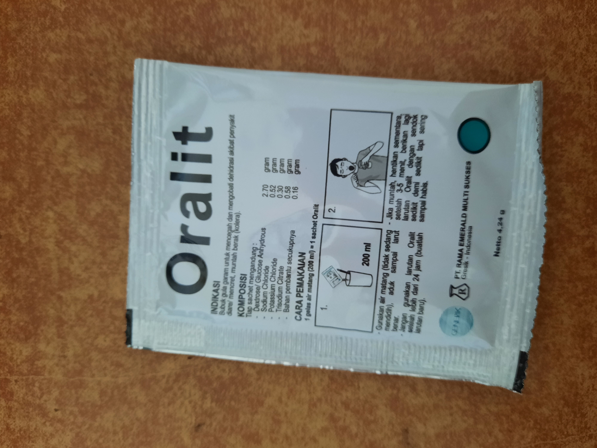 The image shows a white packet of "Oralit", a rehydration salt, lying on a textured brown surface. The packet has black text and some illustrations. The text indicates the product's purpose ("INDİKASI"), composition ("KOMPOSISI"), and instructions for use ("CARA PEMAKAIAN"). There is an illustration of dissolving the powder in water and another of a person drinking the solution. The packet also displays the manufacturer's name "PT. RAMA EMERALD MULTI SUKSES" and its location in "Gresik - Indonesia". The net weight of the product is listed as "Netto 4,24 g". The image is taken from a top-down perspective, and the lighting suggests it is indoors. There is no indication of the product's location in the city of Lhokseumawe, Indonesia.