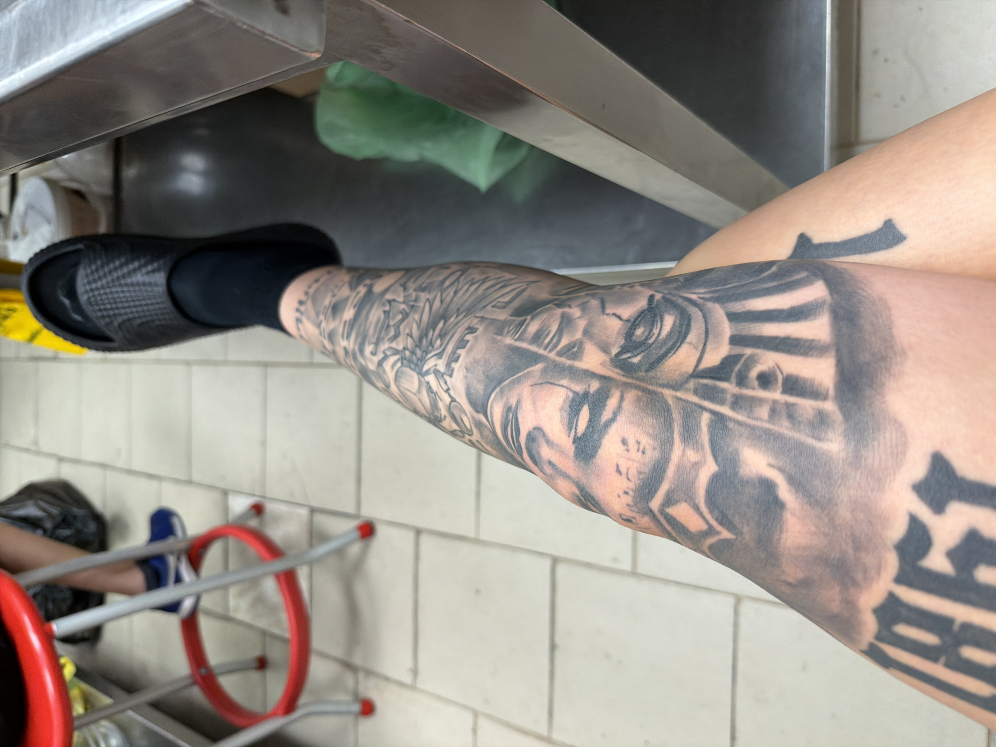 This media file shows a close-up view of a person's leg, covered in intricate black and grey tattoos. The leg is positioned horizontally, suggesting the person may be sitting or lying down. A black slide sandal and a black sock are visible on the foot. The background features a tiled floor and a stainless steel surface, possibly part of a kitchen or workshop. A green plastic bag and some red-rimmed metal objects, resembling part of a rack or stand, are also visible in the background, along with a blue and white athletic shoe. The lighting appears to be indoor, with no clear indication of the time of day. The overall impression is a candid shot focusing on the detailed body art. No visible text is present in the image.