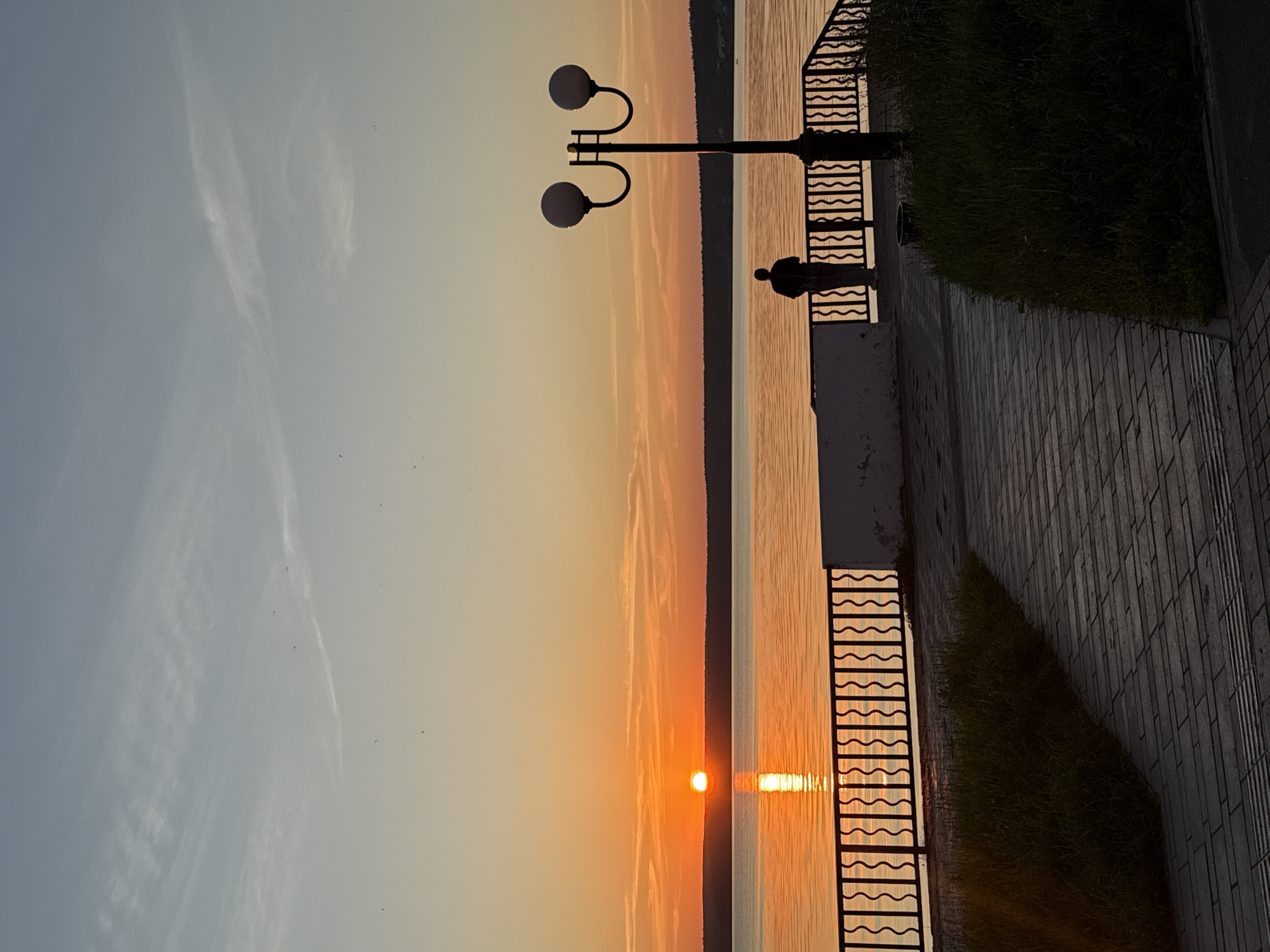 A lone figure stands silhouetted against a sunset over the Votkinsk Reservoir in Votkinsk, Russia. The sun, a bright orange orb, is partially submerged below the horizon, casting a warm glow across the water and the sky. The sky transitions from a pale yellow at the horizon to a soft blue at the top, with faint wispy clouds scattered throughout.
In the foreground, a paved walkway curves towards the reservoir, bordered by a grassy embankment on the left and a decorative black metal railing on the right. The railing runs along the edge of a low, white wall. A two-headed streetlamp stands near the railing, its silhouette also dark against the vibrant sky. The silhouetted figure is positioned on the walkway, facing the sunset and the water, seemingly contemplating the scene. The water is calm, reflecting the colors of the sky and the sunset. The overall atmosphere is one of tranquility and natural beauty.