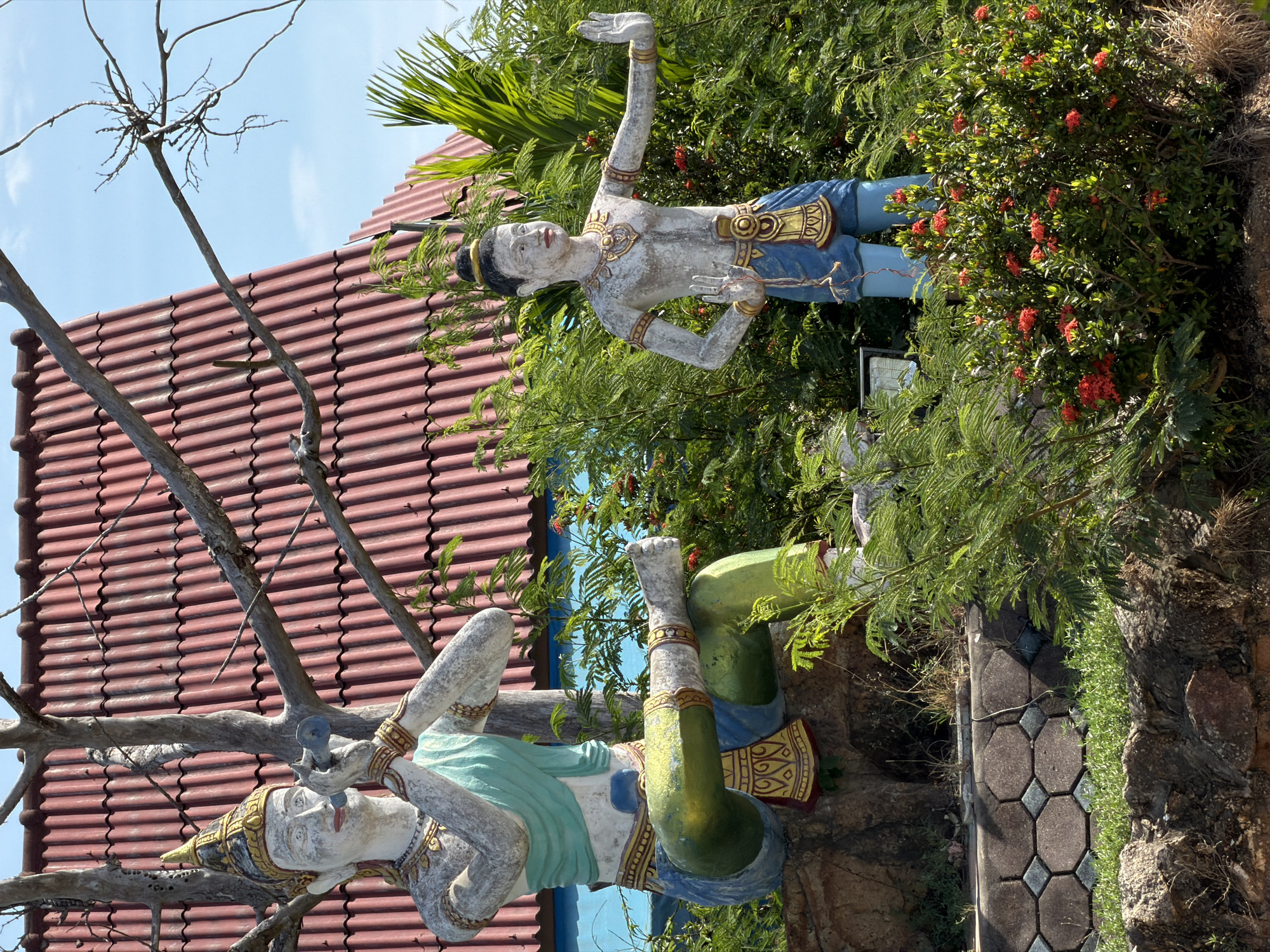 This image depicts a scene at Baan Plai Laem, Thailand, featuring two statues and surrounding vegetation. The statue on the left is a seated figure with a regal headdress, draped in a turquoise cloth, and holding a musical instrument to its lips. It is positioned on a rocky outcrop. The statue on the right is standing, with arms raised in a gesture of greeting or protection. It wears a blue garment and gold adornments. Both statues are weathered and appear to be made of concrete or a similar material. Lush green foliage, including ferns and flowering bushes with red blossoms, surrounds the statues. In the background, a red-tiled roof of a building is visible against a clear blue sky. The scene is bathed in bright sunlight, suggesting a daytime setting.
