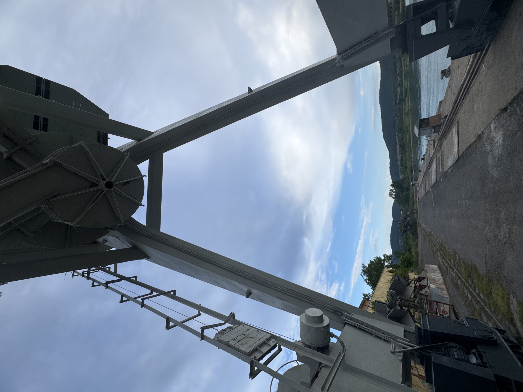 The image depicts a port facility in Burgas, Bulgaria. A large, gray gantry crane dominates the foreground, its metal structure forming an imposing silhouette against a partly cloudy sky. The crane's triangular supports extend downwards, with a ladder or access mechanism visible on the left. Beyond the crane, a paved pathway runs alongside a body of water, likely the Burgas Bay. In the distance, lush green hills form the backdrop. The scene appears to be an industrial area with remnants of previous activity, including what looks like discarded machinery or equipment. The overall atmosphere is one of quiet industrial decay. There are no people visible in the image.