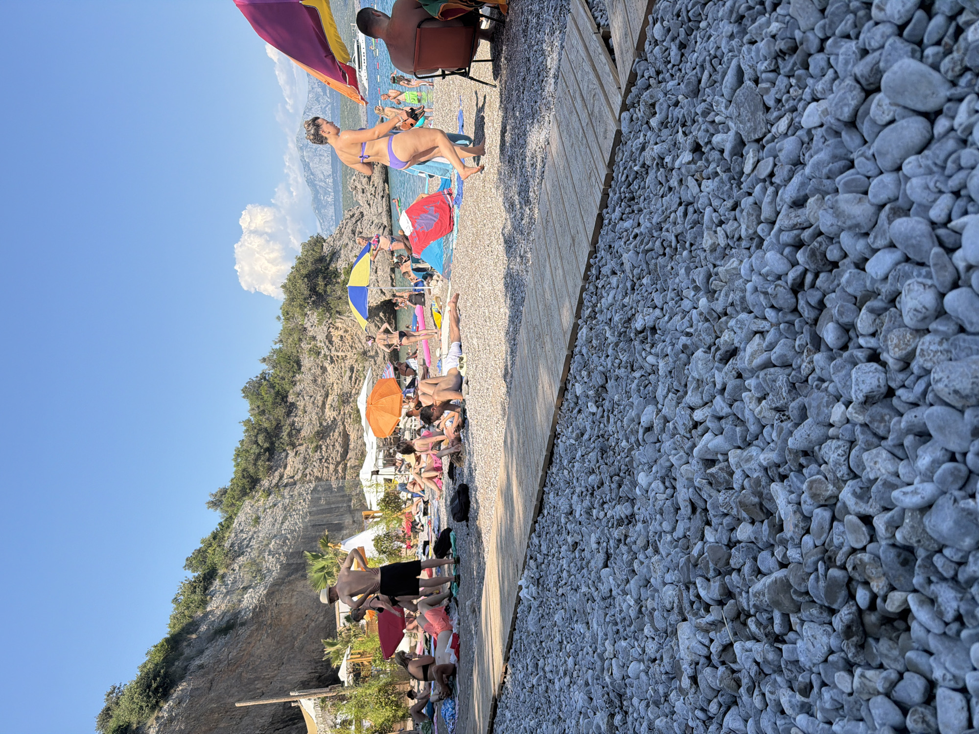 The image depicts a crowded pebble beach on a sunny day, with many people sunbathing and relaxing. In the foreground, a wooden walkway is laid across the pebbles. Several colorful beach umbrellas are visible, providing shade. In the background, a rocky cliff face covered with green vegetation rises behind the beach. The sea is calm and a few boats are anchored in the distance. People are wearing swimwear, and some are lying on towels or beach chairs. The atmosphere is one of leisure and recreation. This scene is consistent with the popular beaches found in the vicinity of Radovići, Montenegro, known for their natural beauty and tourist amenities.