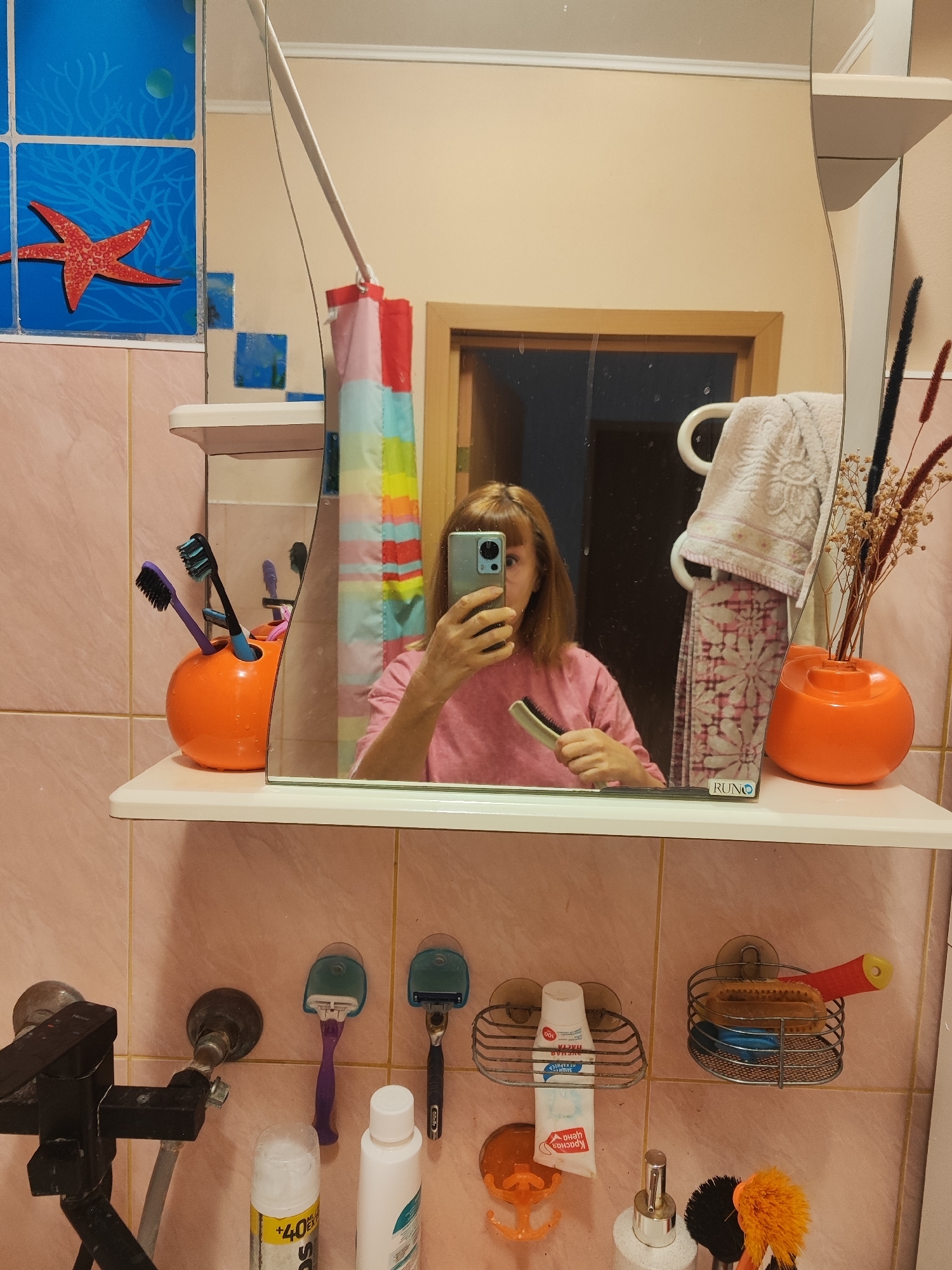 The image depicts a bathroom scene, presumably in an apartment in Gvardeysk, Russia, given the context of the prompt. A woman is taking a selfie in a mirror. She has short, light brown hair and is wearing a pink t-shirt. In her right hand, she holds a mobile phone, and in her left, she is holding a hairbrush.
The bathroom is tiled with light pink tiles. Above the sink area, a mirror reflects the scene. On a shelf below the mirror, several items are visible: an orange toothbrush holder containing several toothbrushes, a vase with dried flowers, and a towel hanging on a rack. To the right of the sink, there are two shavers mounted on the wall, a wire basket holding a bar of soap and a toothbrush, and another wire basket holding a scrub brush. Below the shelf, on the wall, are various plumbing fixtures and bottles of toiletries, including a can of shaving foam and a bottle of liquid. The overall impression is of a functional, lived-in bathroom.