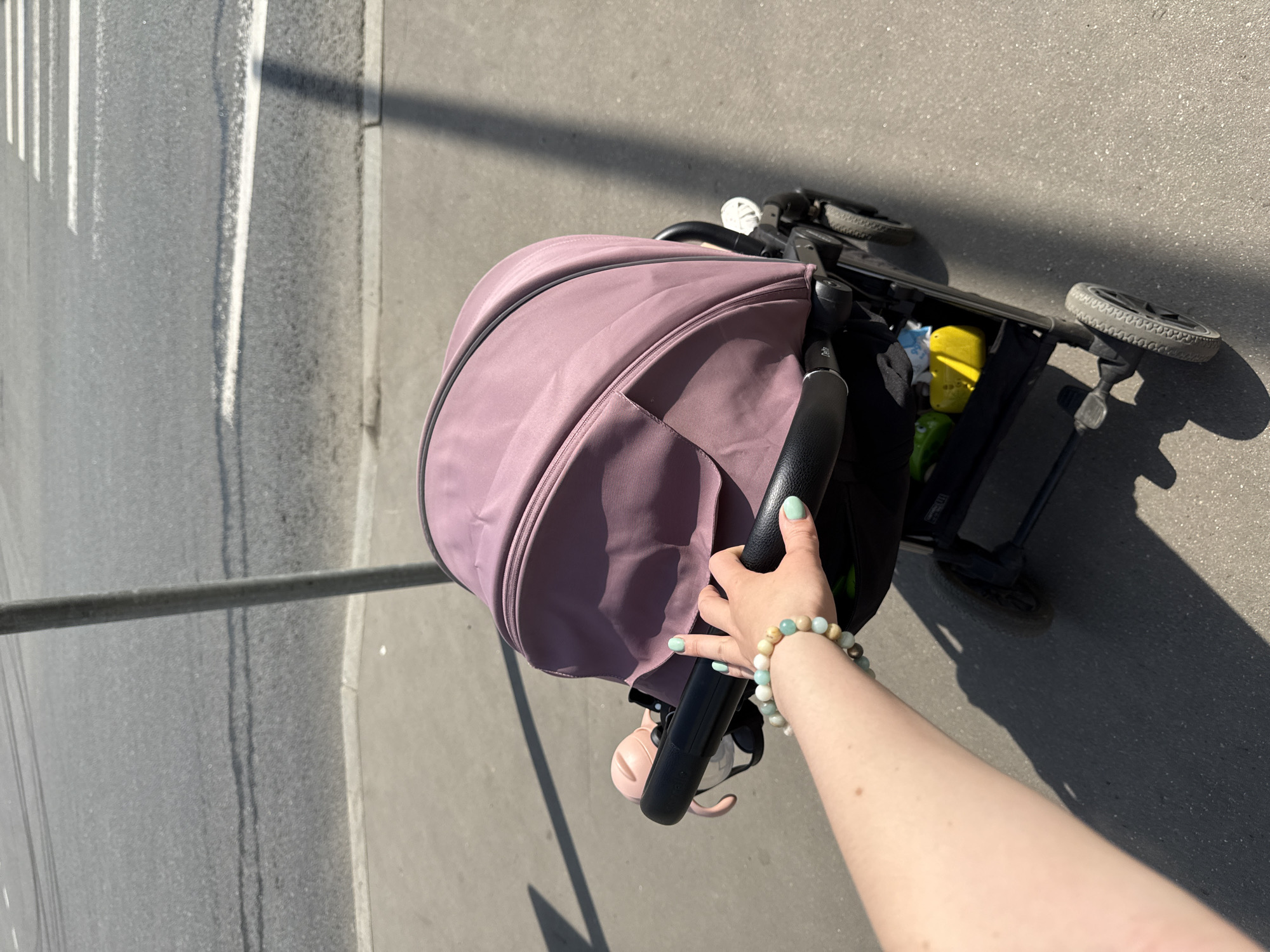 The image is taken from a high angle perspective, showing a person's arm and hand pushing a pram on a street. The pram has a mauve-colored canopy, and a black frame. The person is wearing a bracelet. The location appears to be a sidewalk adjacent to a street, possibly in a city in Moscow, Russia, based on the style of street markings.