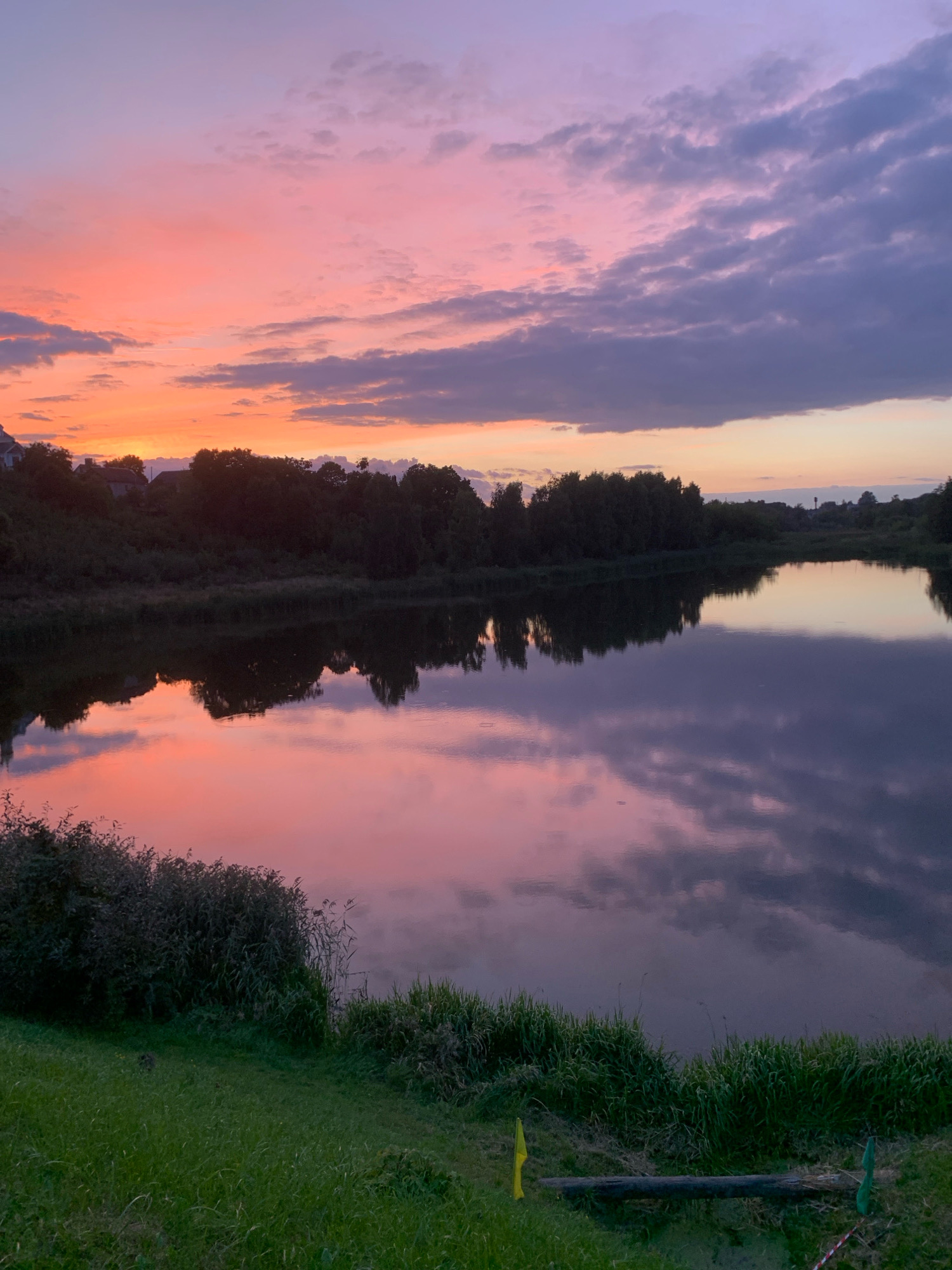The image depicts a sunset over a tranquil body of water, likely a lake or river, in Kapyĺ, Belarus. The sky is a gradient of soft pinks, oranges, and purples, with scattered clouds adding texture and depth. The vibrant colors of the sunset are mirrored in the calm surface of the water, creating a surreal and peaceful reflection.
Along the far bank of the water, a dense line of trees forms a dark silhouette against the colorful sky. In the distance, hints of human habitation can be seen with the roofs of houses peeking through the trees on the left side of the image.
The foreground of the image is dominated by lush green grass, sloping down towards the water's edge. Some bushes and reeds grow along the bank, adding to the natural setting. In the bottom right quadrant, a rustic wooden log lies horizontally across the grass, with small yellow and green flags placed on either side. This suggests a possible recreational area or a marker of some kind. The overall atmosphere is one of natural beauty and serene twilight.
