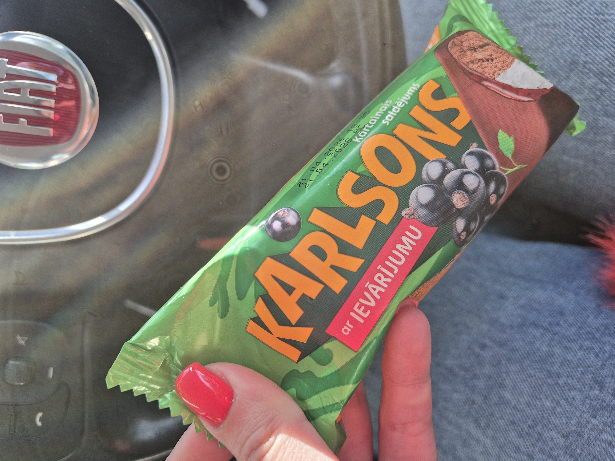 A hand with red nails holds a green Karlsons ar Ievarijumu black currant ice cream bar in front of a Fiat steering wheel and blue jeans. The image appears to be taken indoors, possibly inside the car. The expiration date on the bar is visible as 11.04.2023. The scene does not provide sufficient information to determine a specific location within the Breckland District or even the United Kingdom.