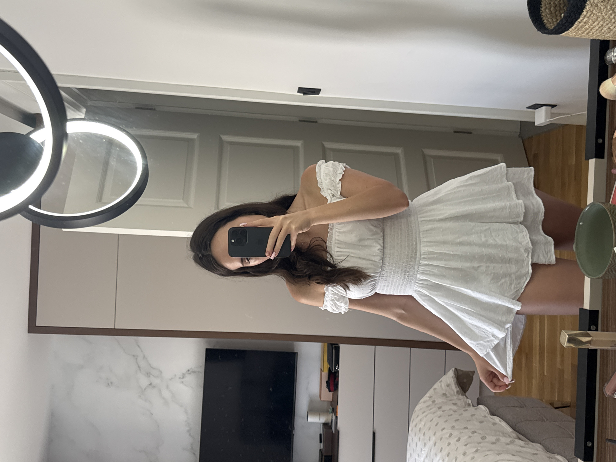 A young woman stands in a bedroom, taking a mirror selfie. She is wearing an off-the-shoulder white mini dress with a smocked bodice and a flared skirt. Her dark hair falls around her shoulders, and she holds a black smartphone in her right hand. To her left is a bed with a grey headboard and white patterned bedding. Behind her, a white dresser is visible, topped with various items including a black television. A large mirror reflects the scene, as well as a modern light fixture hanging from the ceiling. The room appears to be neutrally decorated with white walls and doors.