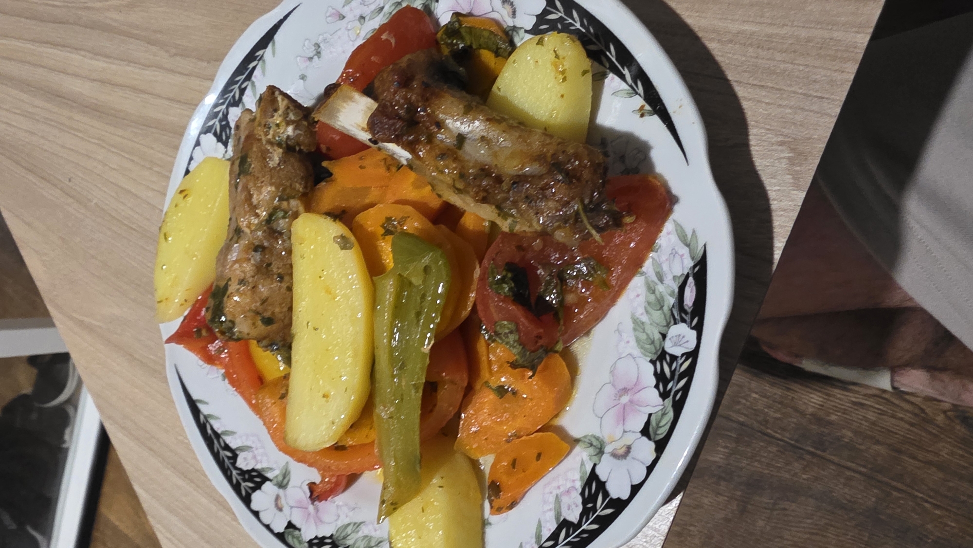 A close-up, overhead shot shows a plate of roasted chicken, potatoes, and bell peppers on a wooden table. The plate has a floral pattern around the rim. The food appears to be seasoned with herbs and spices. In the background, a blurry image of shoes on a shelf is visible. The lower portion of the image shows a person's legs and clothing.