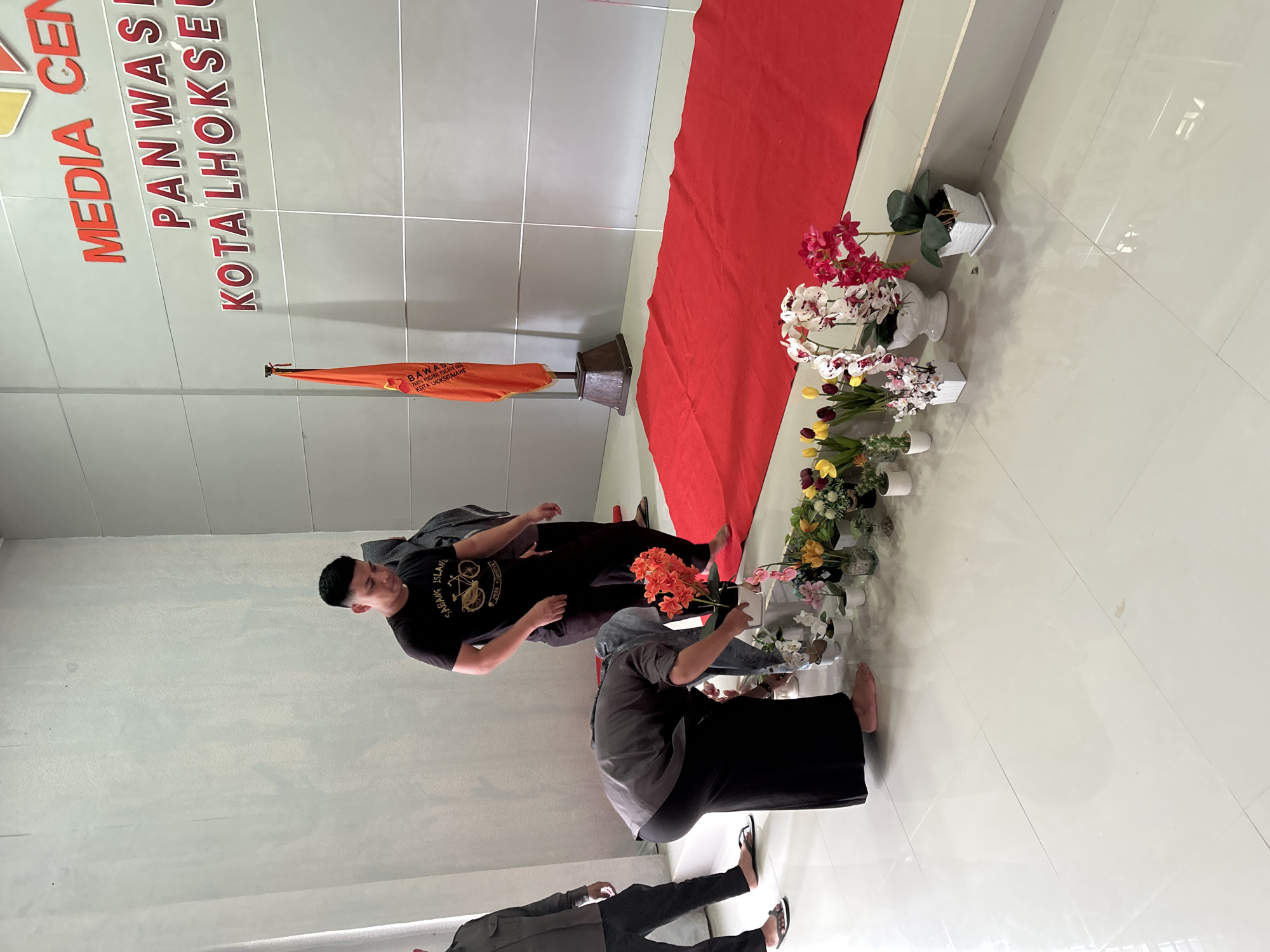 The image shows a scene inside a building in Lhokseumawe, Indonesia. The text "MEDIA CEN PANWAS KOTALHOKSEL KOTA LHOKSEUMA WE PARTA PENGARAS PORLAR BAWAS" is visible on the wall. An orange flag and a red runner are present in the background, and several potted flowers line the side of the runner. Three people are present. One man is wearing a black t-shirt with a bicycle design. A woman in a headscarf is arranging orange flowers in a pot. Another person is partially visible at the bottom of the frame. The room has a glossy tiled floor.