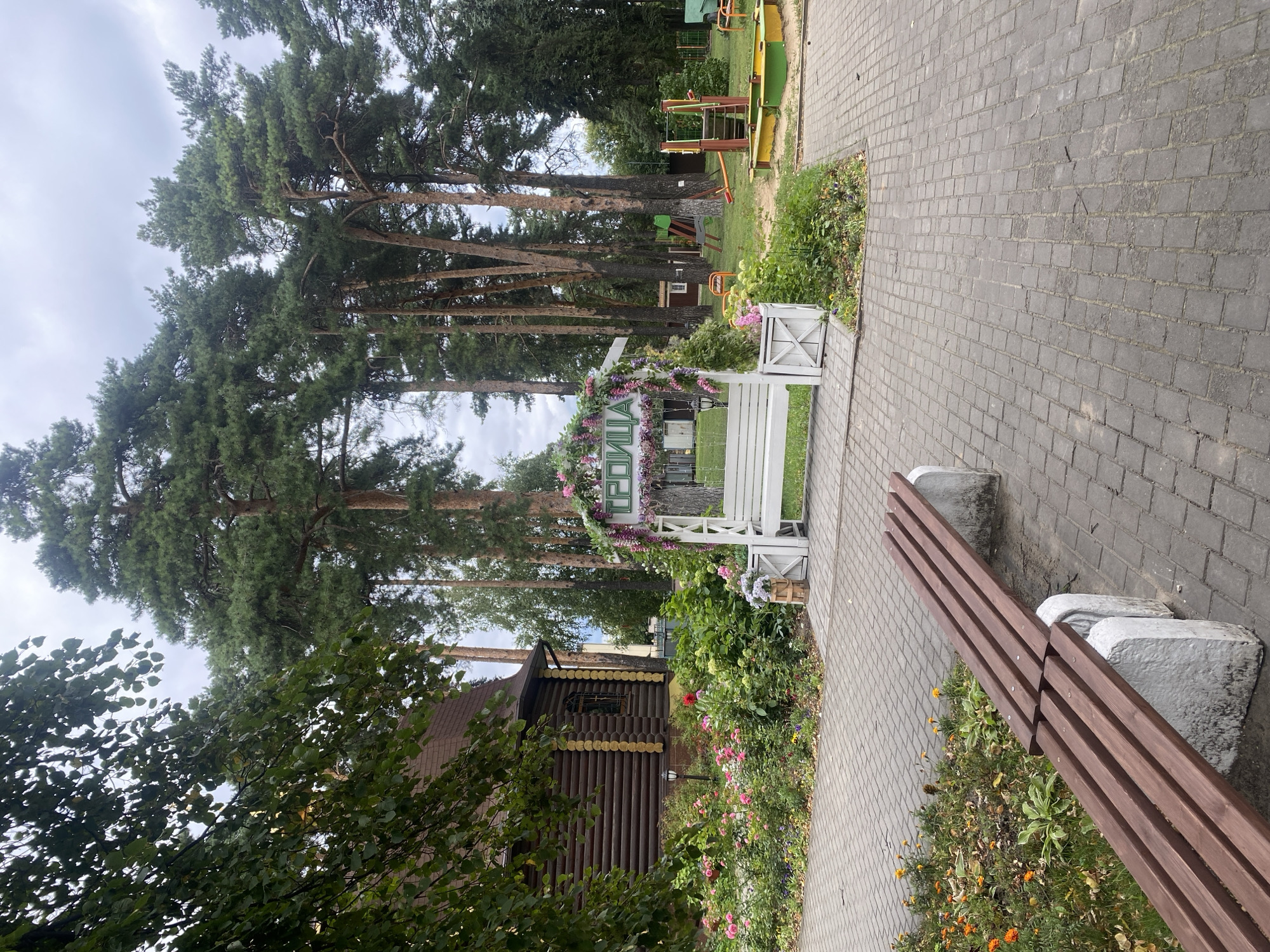 A wide-angle, eye-level shot depicts an outdoor scene with a prominent white archway adorned with flowers and greenery. The archway features the Cyrillic word "ТРОИЦА" (Troitsa) in green lettering. A white bench sits within the archway. To the left of the archway is a log building with a brown roof and a detailed facade. A flower bed with various colored flowers, predominantly pink and red, is situated in front of the building and extends towards the archway. A paved walkway, made of grey interlocking stones, leads through the scene. In the foreground, a long wooden bench with three brown planks, supported by two light-colored stone bases, is positioned on the walkway. Behind the archway, a children's playground is visible with colorful structures, including a sandbox and swings, set on a grassy area. Tall pine trees form a dense background, and the sky above is overcast. The overall scene suggests a well-maintained park or recreational area, likely in a town or city like Oktyabrskiy, Russia.