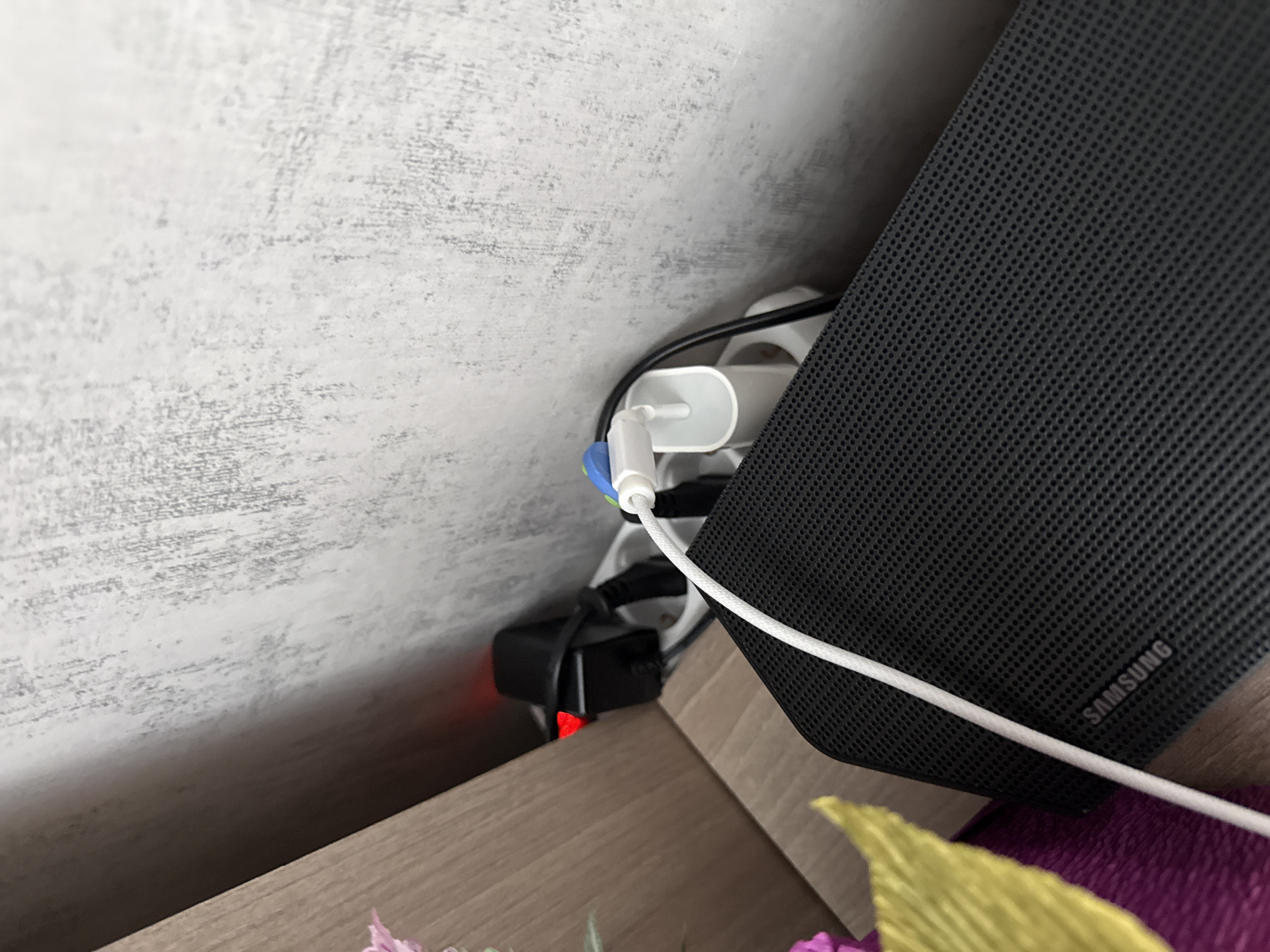 A close-up shot shows a power strip with multiple plugs connected, positioned next to a dark gray Samsung soundbar. The power strip is white, with a black plug and a blue cable wrap visible. A white cable runs from one of the plugs to the soundbar. The soundbar is textured with small perforations and the "SAMSUNG" logo is visible on its side. The power strip and soundbar are placed on a wooden surface, with part of a floral decoration visible at the bottom. The wall behind the devices has a light gray, textured finish. The image appears to be taken indoors, and there's no clear indication of a specific location in Lovech, Bulgaria.