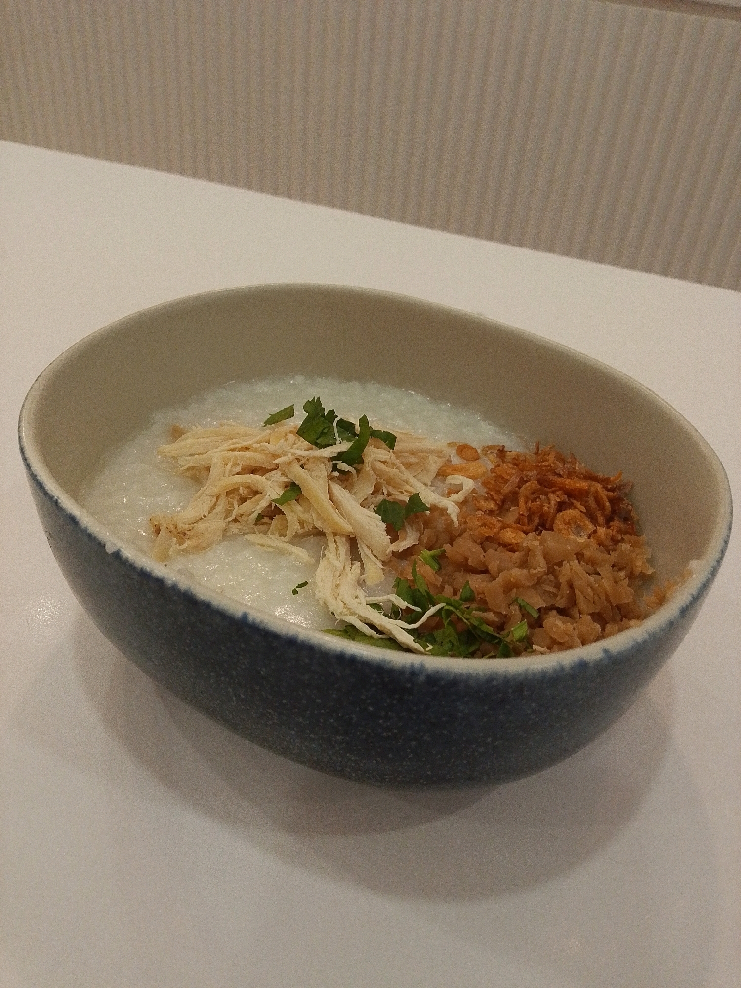 A close-up, eye-level shot showcases a bowl of congee, likely served in Semarang, Indonesia. The congee is topped with shredded chicken, fried shallots, and chopped parsley. The bowl itself is a rustic ceramic with a speckled blue exterior and a lighter colored interior. It sits on a plain white surface, with a textured, vertically striped background visible in the upper portion of the image. The lighting appears to be soft and diffused, creating subtle shadows on the surface.