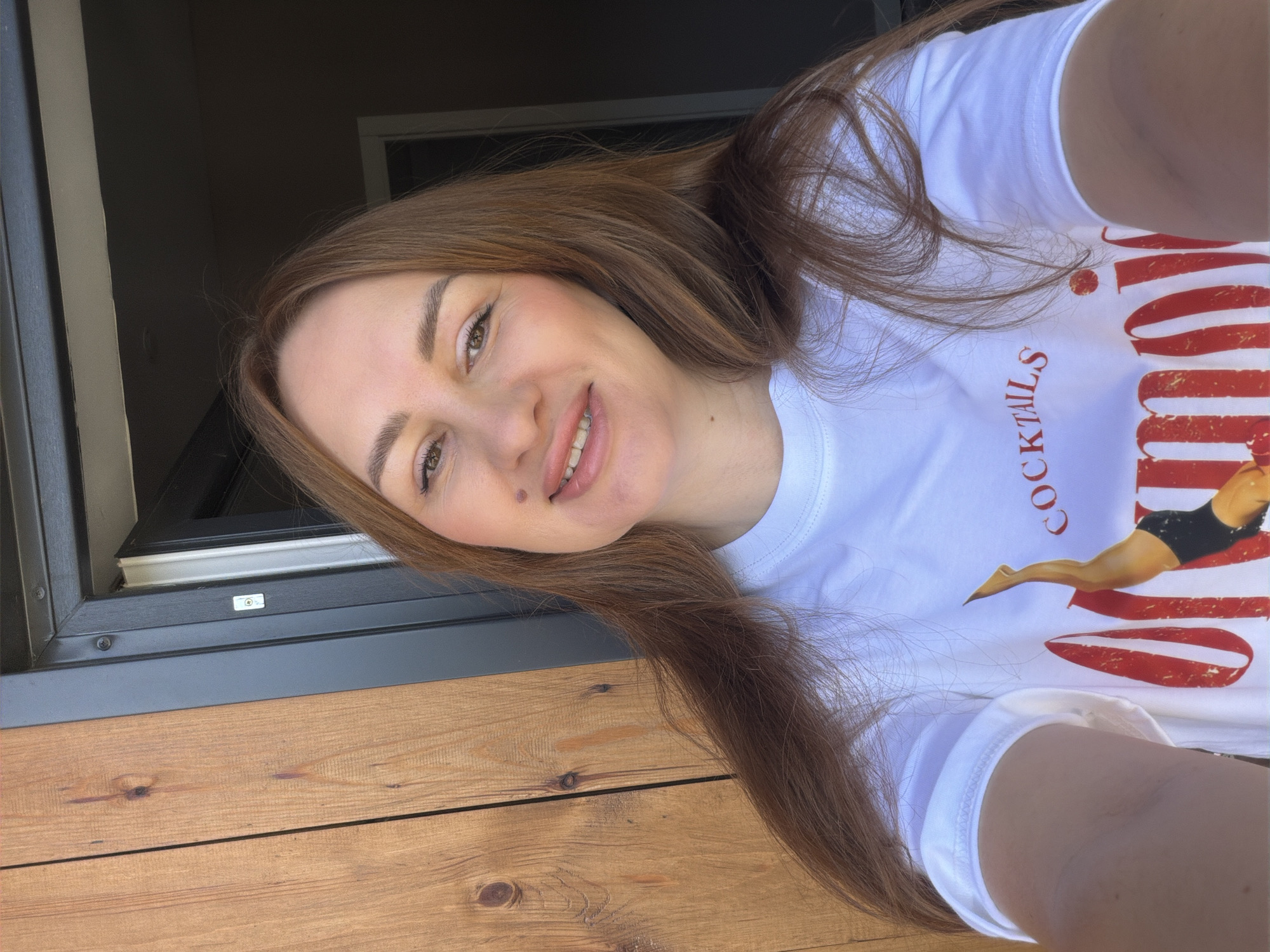 The image shows a woman in her late 20s or early 30s with shoulder-length auburn hair, wearing a white t-shirt with a graphic print. She is outdoors, with a wooden-paneled wall to her left and an open doorway behind her, leading to a dark interior. The lighting suggests it is daytime. The woman is looking directly at the camera and smiling slightly. The graphic on her shirt appears to be red lettering that says "COCKTAILS" and "Manoir" with an image of a person in a diving pose. The overall scene does not provide any specific clues to pinpoint its location within Malinovka, Russia, or any other city.