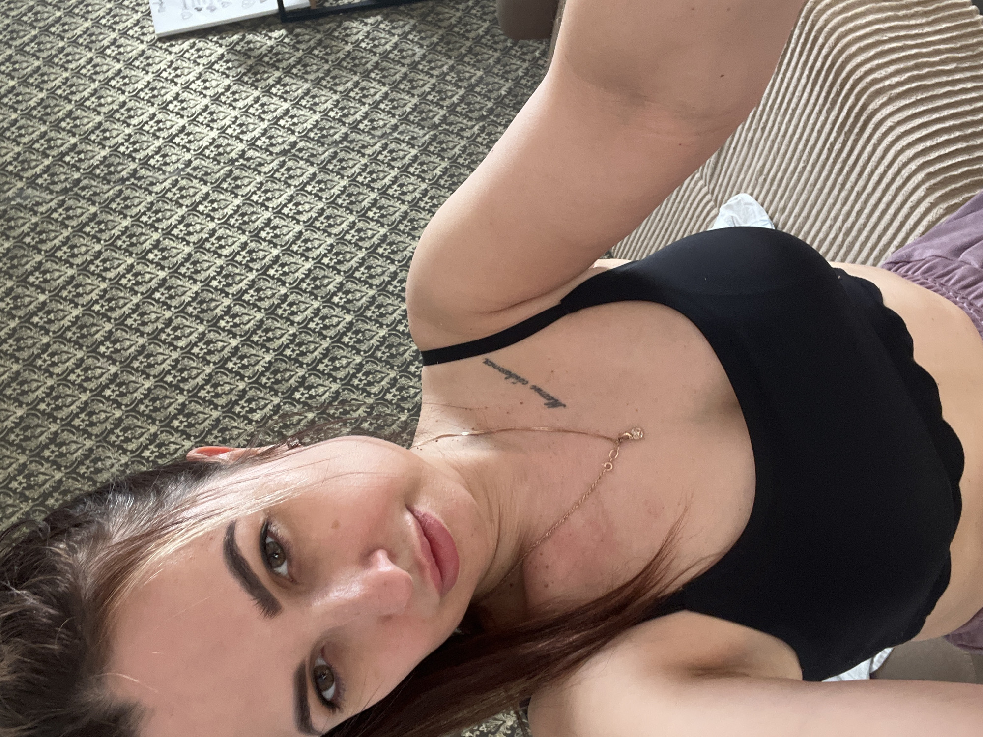 A selfie of a woman lying down, with her head at the bottom left of the frame and her torso at the top right. She is wearing a black sports bra and a gold necklace. A tattoo is visible on her collarbone. The background includes a patterned floor and a striped sofa or bed. The location and city are unknown.