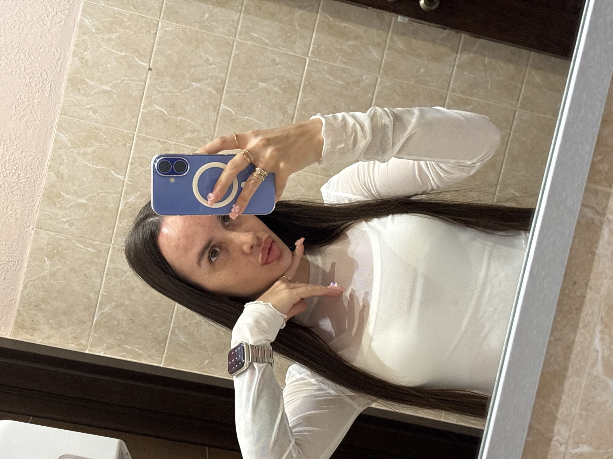 A young woman with long dark hair takes a mirror selfie in a bathroom. She is wearing a white sheer long-sleeved top over a white tank top. She has on rings on her fingers and a smartwatch on her left wrist. The bathroom has tiled walls in a neutral beige color.