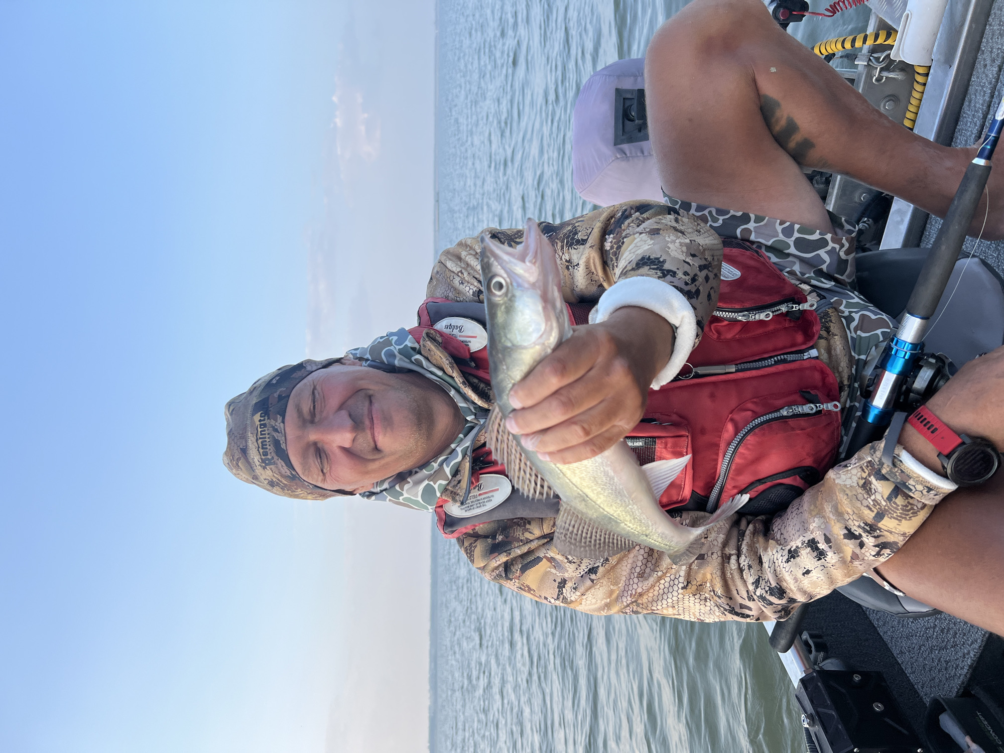 A man is sitting in a boat on a body of water, holding a small fish. He is wearing a camouflage beanie, a camouflage jacket, and a red life vest. The fish is light green with silver scales and its mouth is open. The man is smiling with his eyes closed. The water is calm and the sky is clear blue with a few wispy clouds. The background shows a faint shoreline. The boat appears to be on a lake or a large river, consistent with the environment of Novopichugovo, Russia.