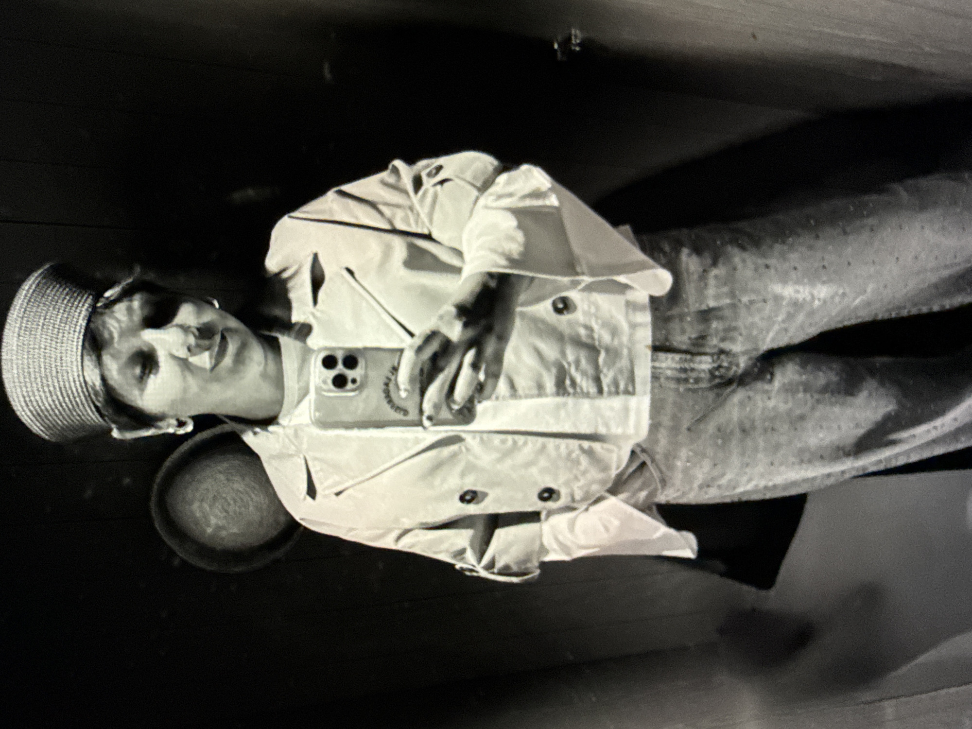 A person is standing in a room holding a smartphone. They are wearing a hat, a light-colored jacket with buttons, and pants. A circular object is located near the person's head. The environment is dark. The city of North Kuta, Indonesia, is not visible in the image.