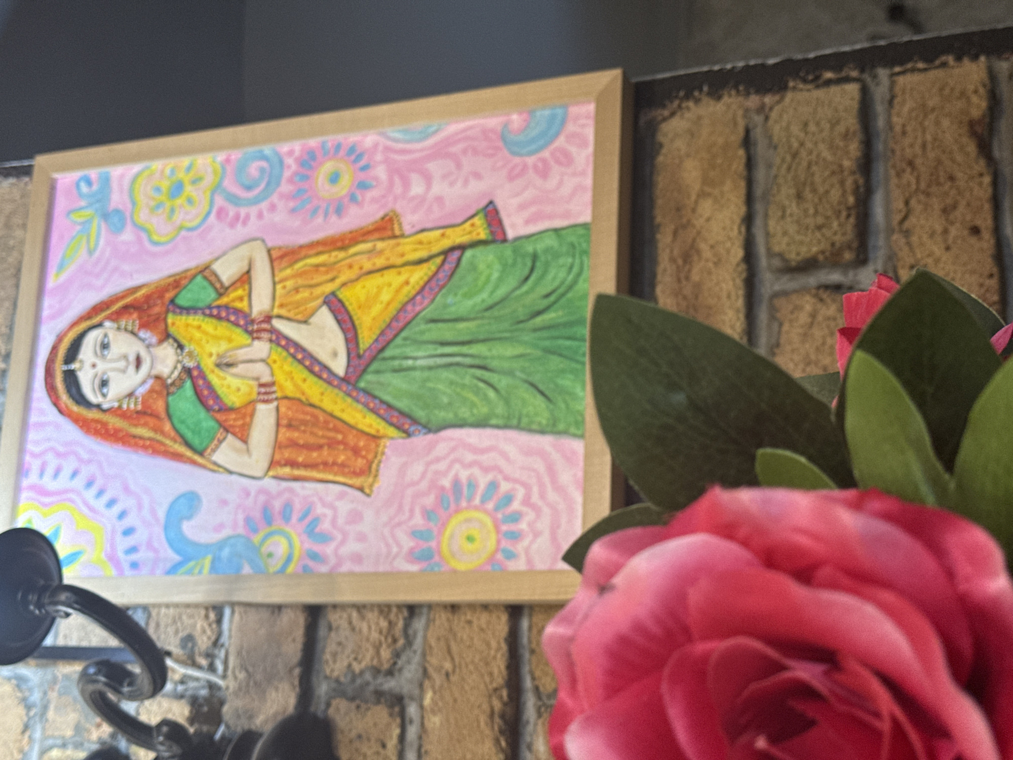 This is a photograph of a painting of a woman in traditional Indian attire, hands pressed together in a gesture of greeting, possibly a greeting or prayer. The painting is framed in a light-colored wooden frame and hangs on a brick wall. In the foreground, out of focus, is a vibrant pink rose with green leaves. The painting features a woman with dark hair, wearing a yellow and green sari with an orange veil. The background of the painting is a soft pink with decorative blue and yellow floral and paisley motifs. The brick wall behind the painting is visible, with visible mortar lines. A dark metal fixture, possibly a lamp holder or decorative element, is visible in the upper left corner. The overall impression is of a decorative piece displayed in a domestic or commercial setting. There is no information within the image to suggest a location in the city of Zagreb, Croatia.