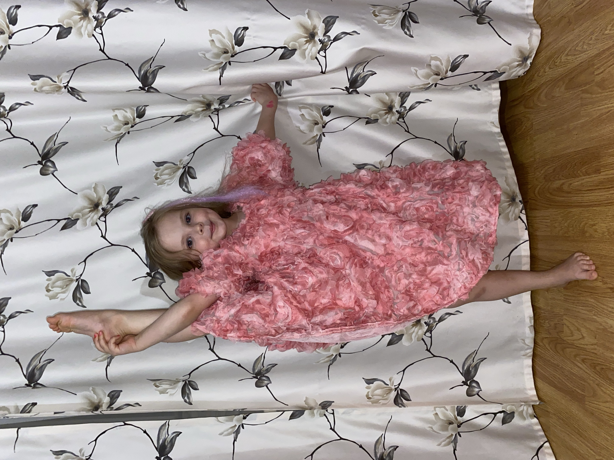 A young blonde girl, wearing a voluminous pink dress made of ruffled or rosette fabric, lies on a white patterned surface while performing a full leg split. She looks directly at the camera with a bright, joyful smile. Her left hand is stretched upwards, grasping the floral patterned fabric, which features light-colored magnolia-like flowers with dark stems and leaves. Her right hand is holding her left foot, pulling it towards her body, further emphasizing her flexibility. A small red heart is visible drawn on the back of her left hand, and faint purple strands are present in her hair. Part of a light brown wooden floor is visible on the right side of the image, suggesting an indoor setting. The scene captures a playful and confident moment in Votkinsk, Russia.
