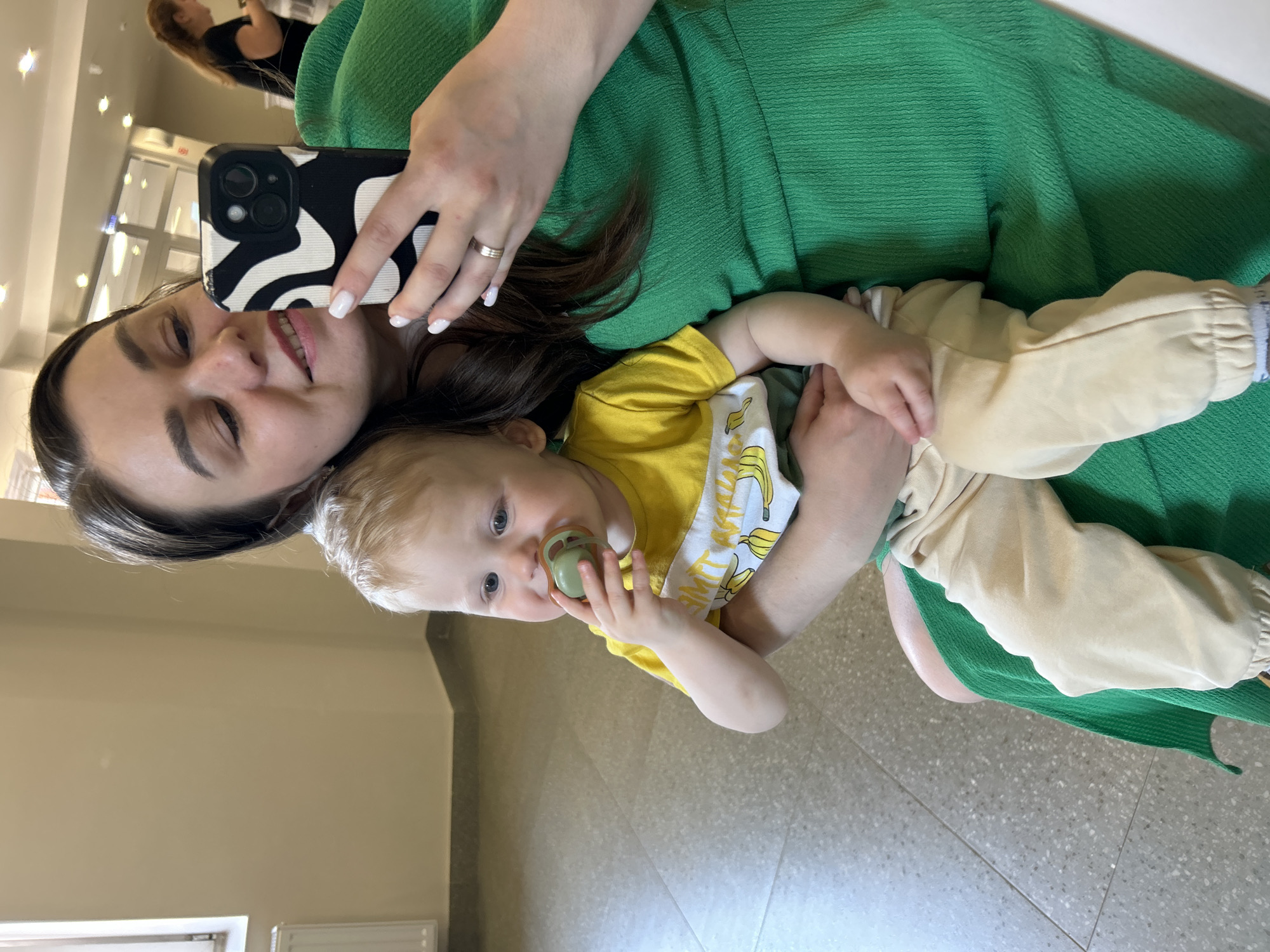 A woman is holding a baby in her arms while taking a selfie. The woman is wearing a green dress and has dark hair. She is smiling at the camera, holding a phone with a black and white patterned case. The baby is wearing a yellow t-shirt with bananas printed on it and light-colored pants. The baby has blonde hair and is holding a pacifier in his mouth. The background appears to be an indoor setting with a shiny floor. It is unclear from the image whether this location is in Izhevsk, Russia or any other city.