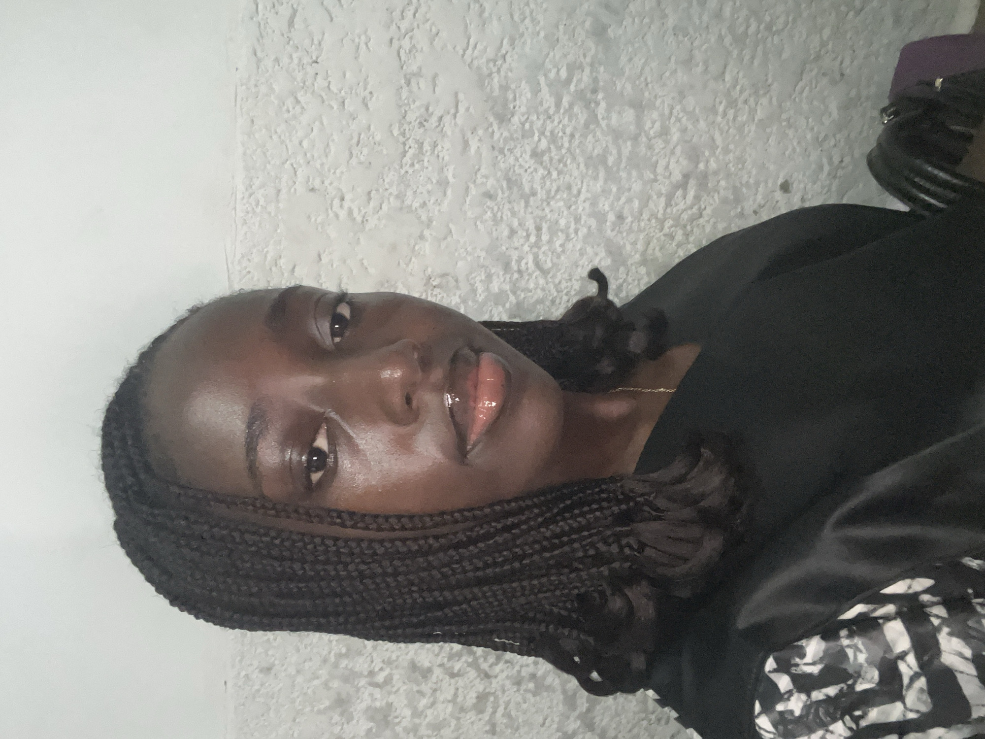 A close-up portrait of a Black woman with braided hair and glossy lips. She is wearing a dark-colored top. The background is a textured, light-colored wall. The location is presumed to be inside a building in the city of Abuja, Nigeria, based on the query, but there are no visual cues to confirm the location or its exact urban context.
